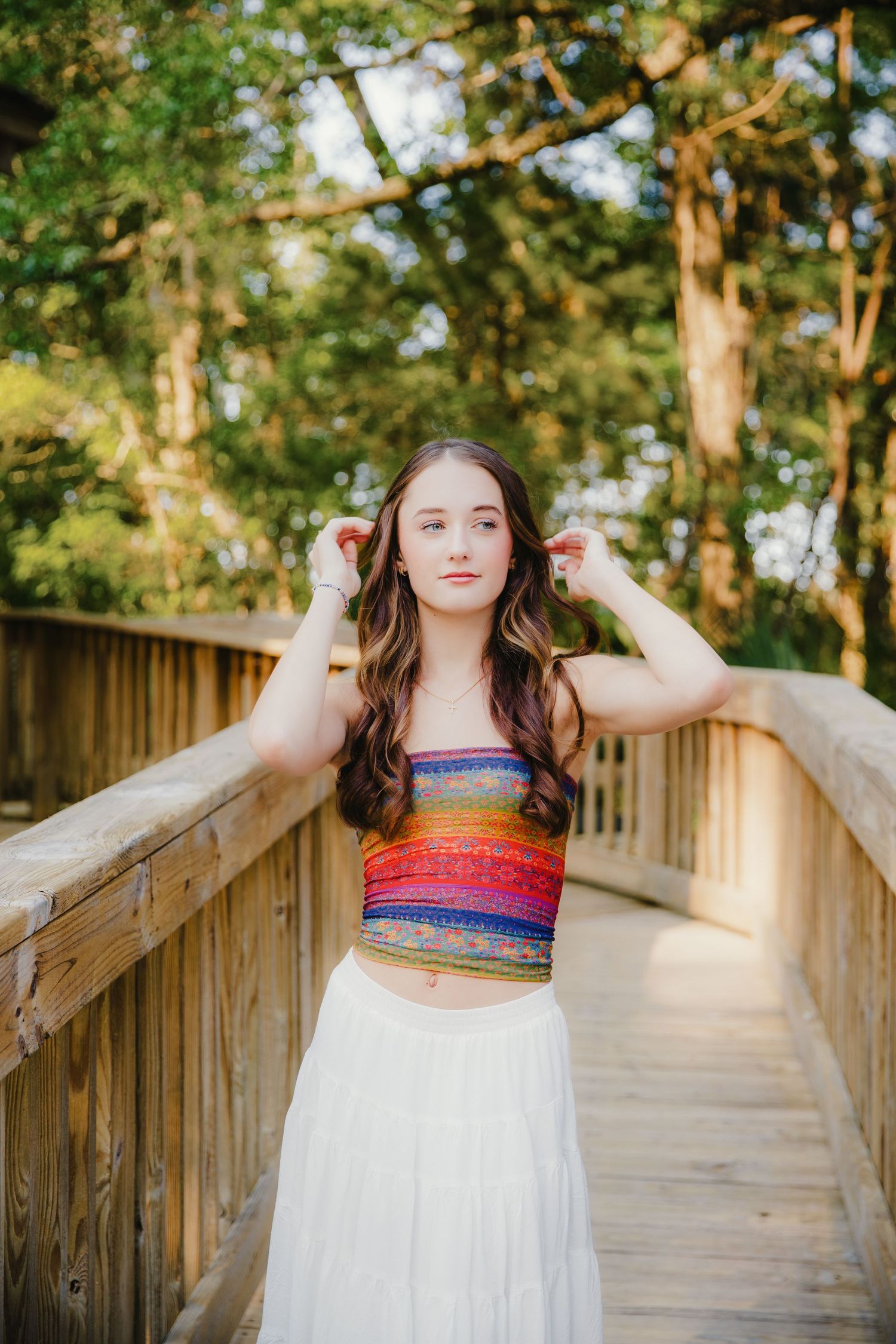 Person in striped top and white skirt standing on wooden walkway among trees.
