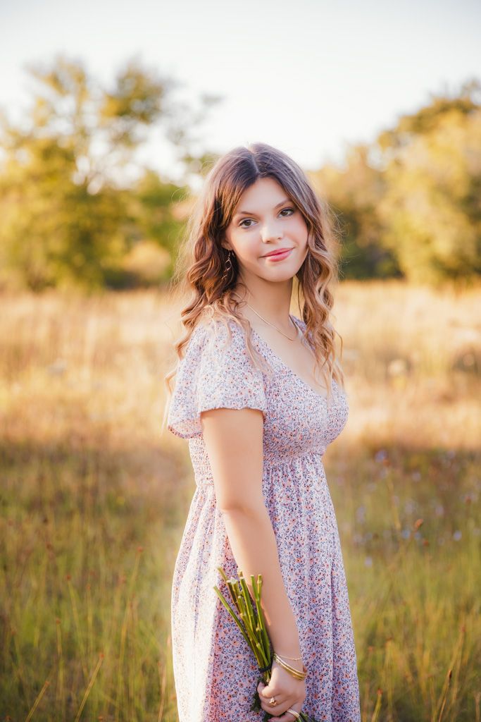 A person in a floral dress stands in a golden field during sunset holding wildflowers.