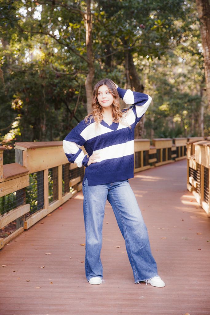 A person wearing a striped navy and white sweater and wide-leg jeans poses on a wooden boardwalk in a forest setting.