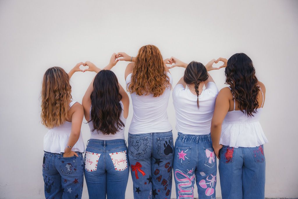 Five women in white tops and decorated jeans stand with their backs to camera showing off artistic denim designs.