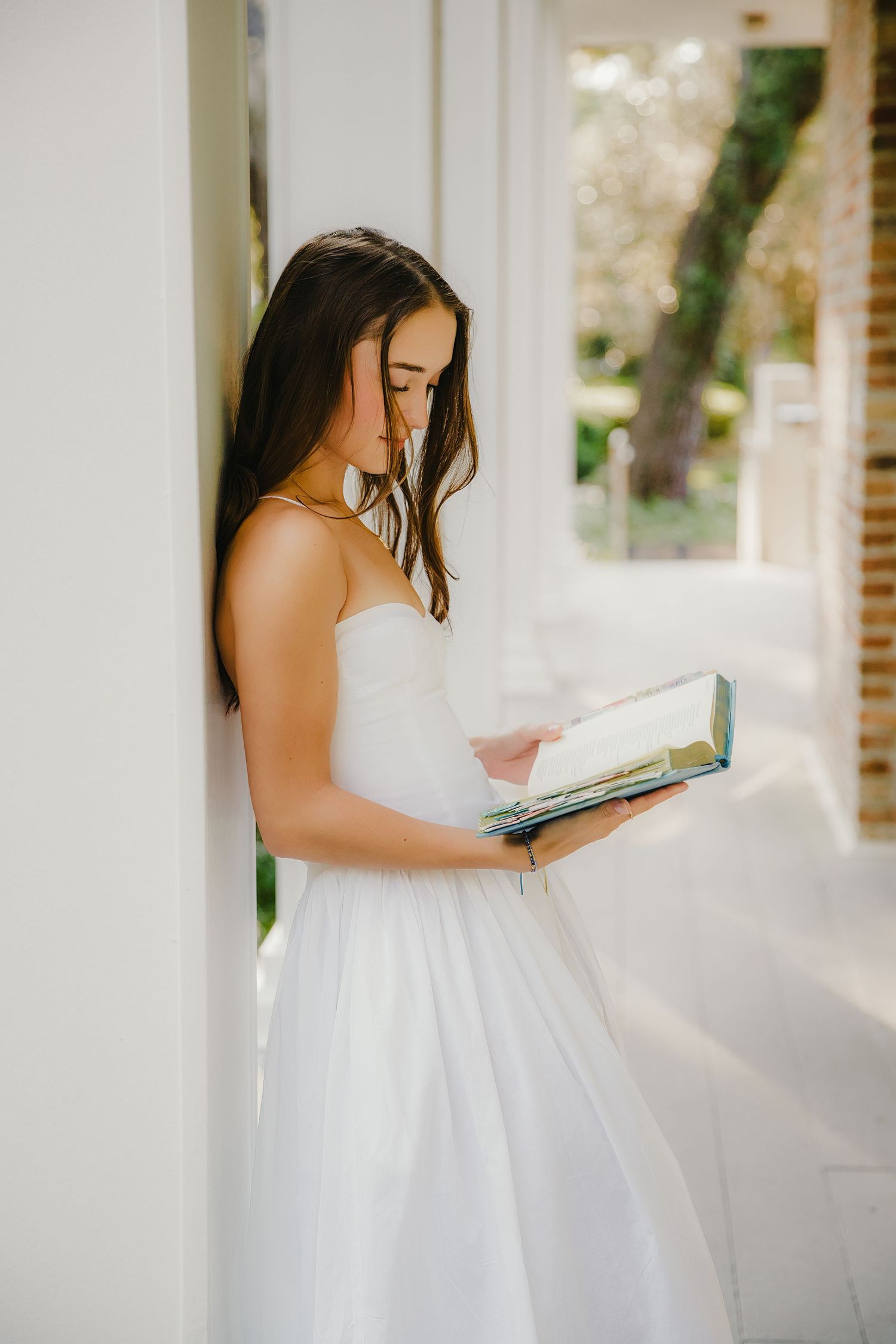 Person in white dress reading book while leaning against white column.