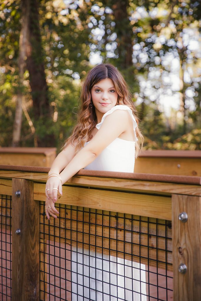 A woman in a white top poses thoughtfully on a wooden deck railing surrounded by sunlit trees.