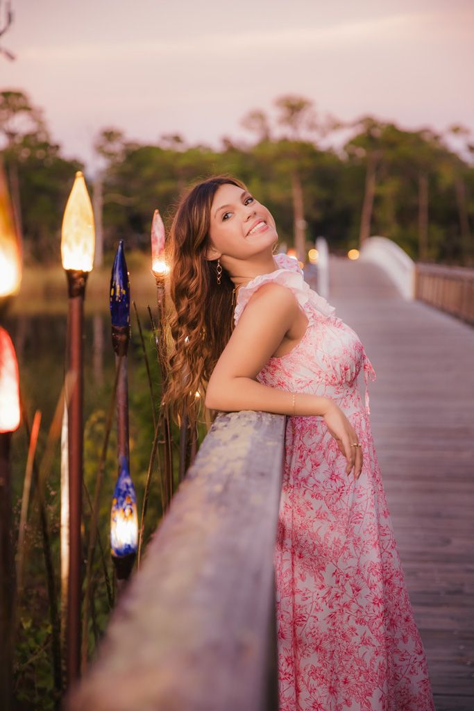 A person in a pink floral dress twirls joyfully on a wooden boardwalk at sunset with tiki torches glowing in the background.