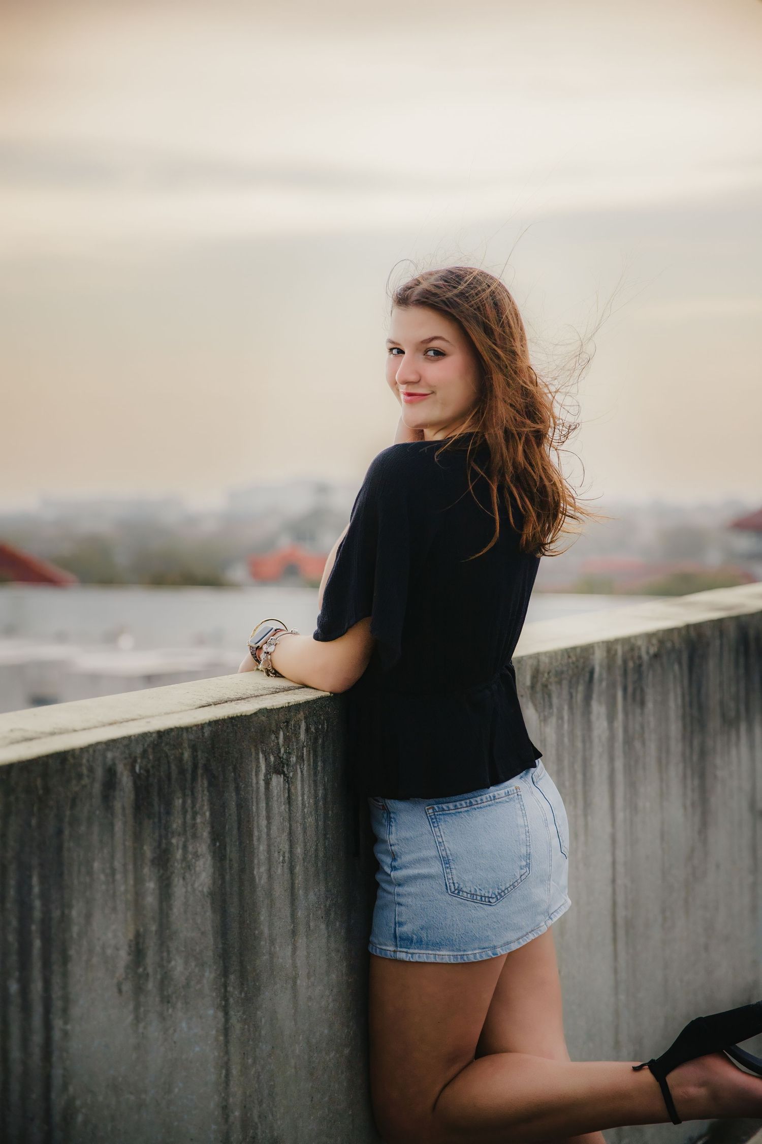 A casual portrait of someone in a black top and denim shorts posing against a concrete wall at sunset.