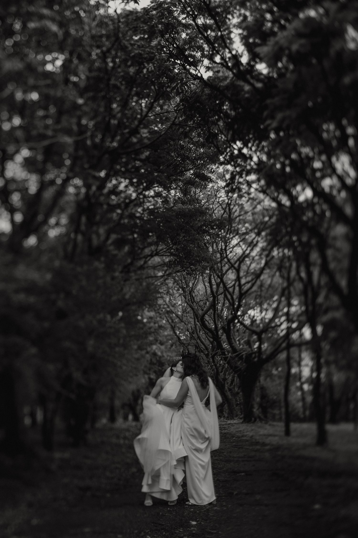 A black and white photograph of two people in flowing white attire walking together on a tree-lined path.