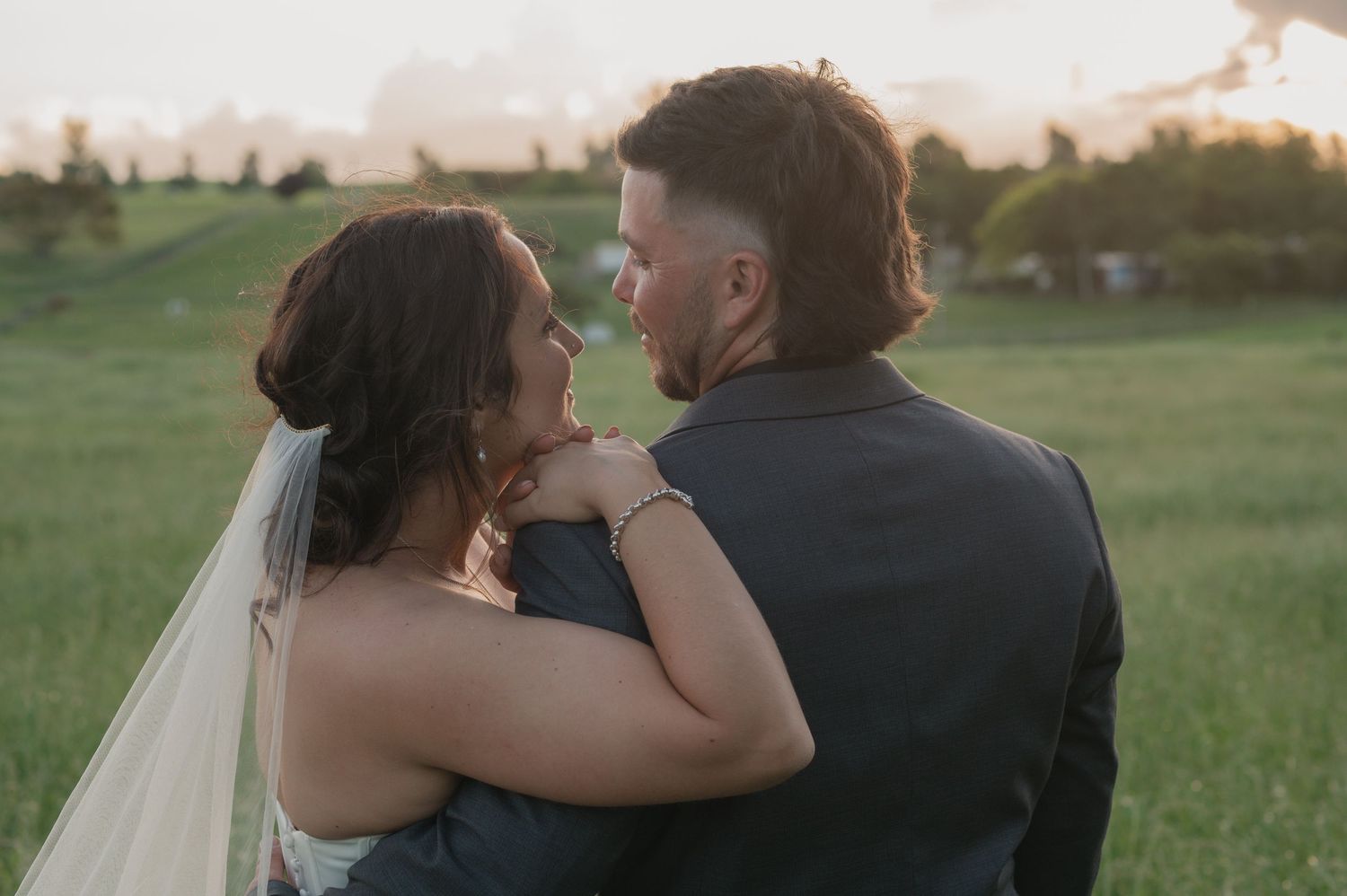 A bride and groom share a romantic moment at sunset in a lush green field, with her veil flowing softly.