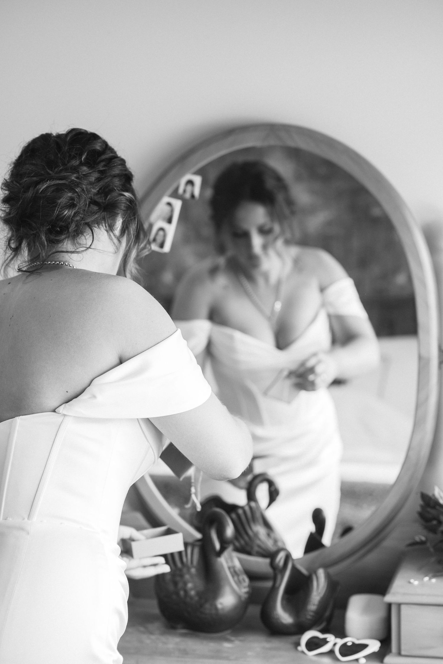 Black and white photo of a bride in a white wedding dress adjusting her gown while looking into a round mirror.