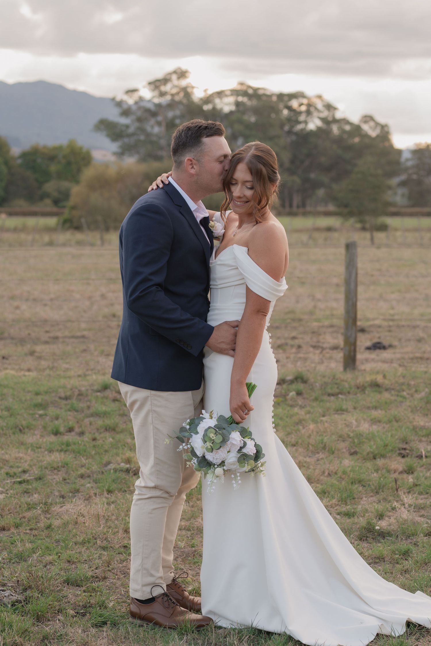 Wedding couple embraces beside Pirongia setting with mountains and eucalyptus trees, bride holding white floral bouquet.