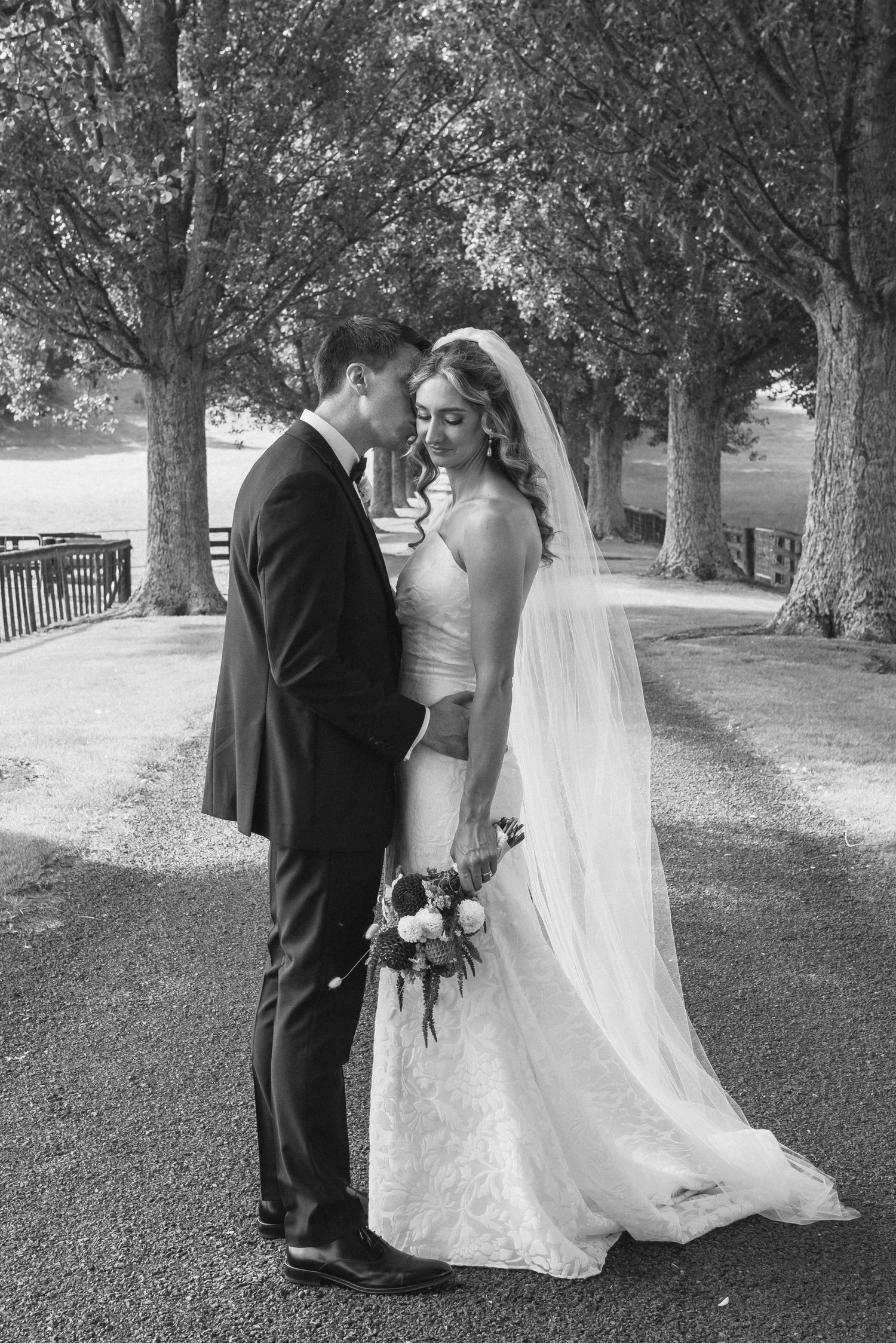 Black and white wedding photo of bride and groom sharing an intimate moment on a tree-lined path, bride holding bouquet.