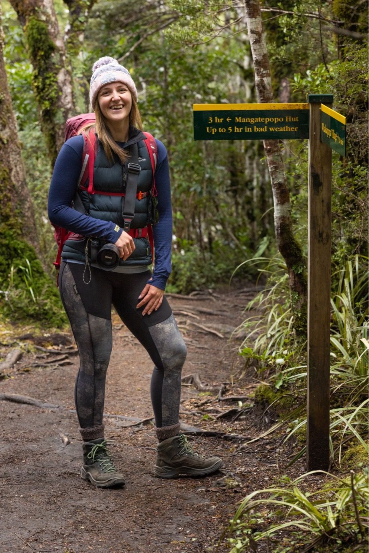 Hiker on forest trail stands next to wooden distance marker post while wearing winter hat, backpack and hiking boots.