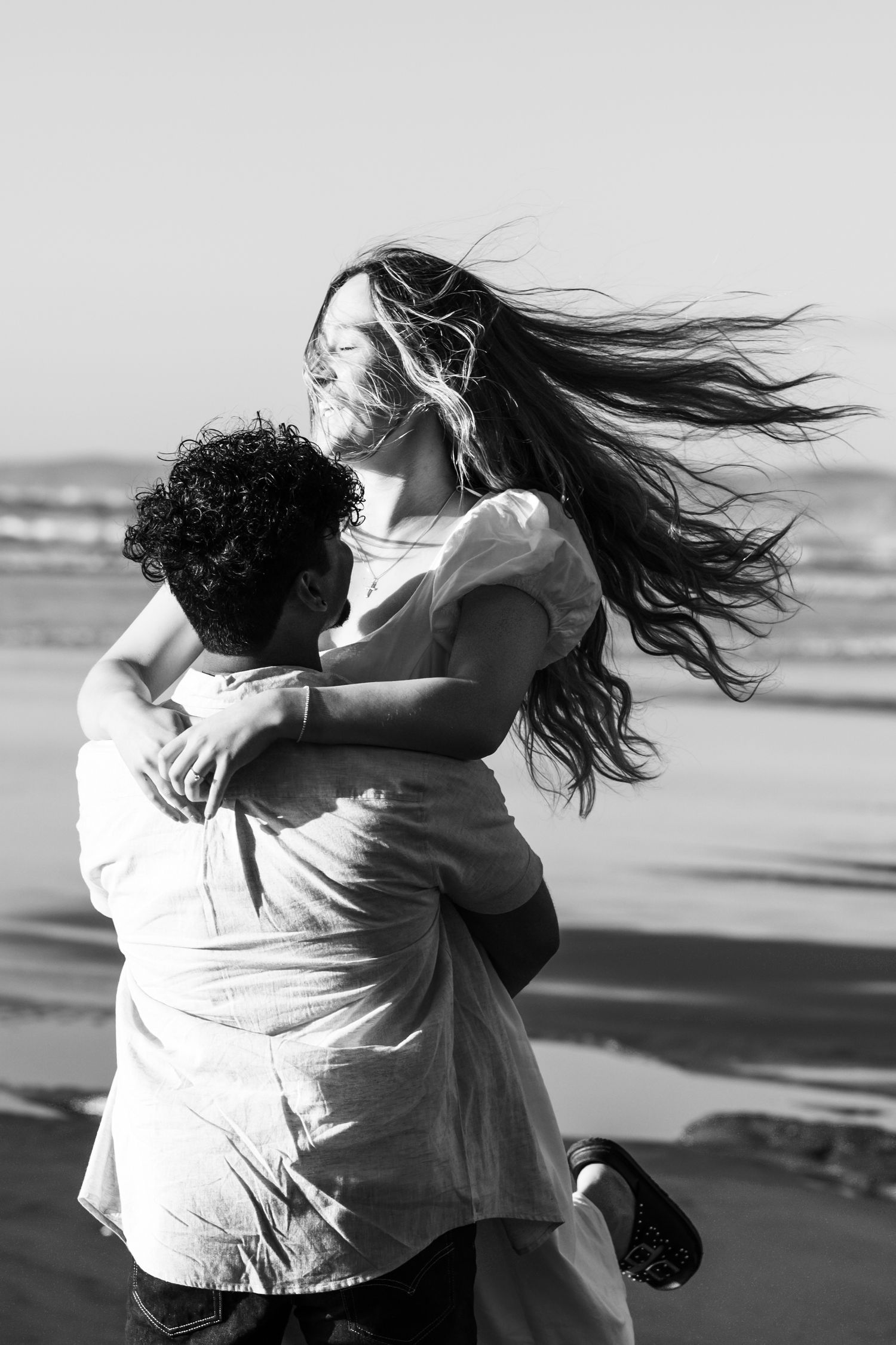 Black and white portrait of couple embracing on windy beach with flowing hair Raglan New Zealand