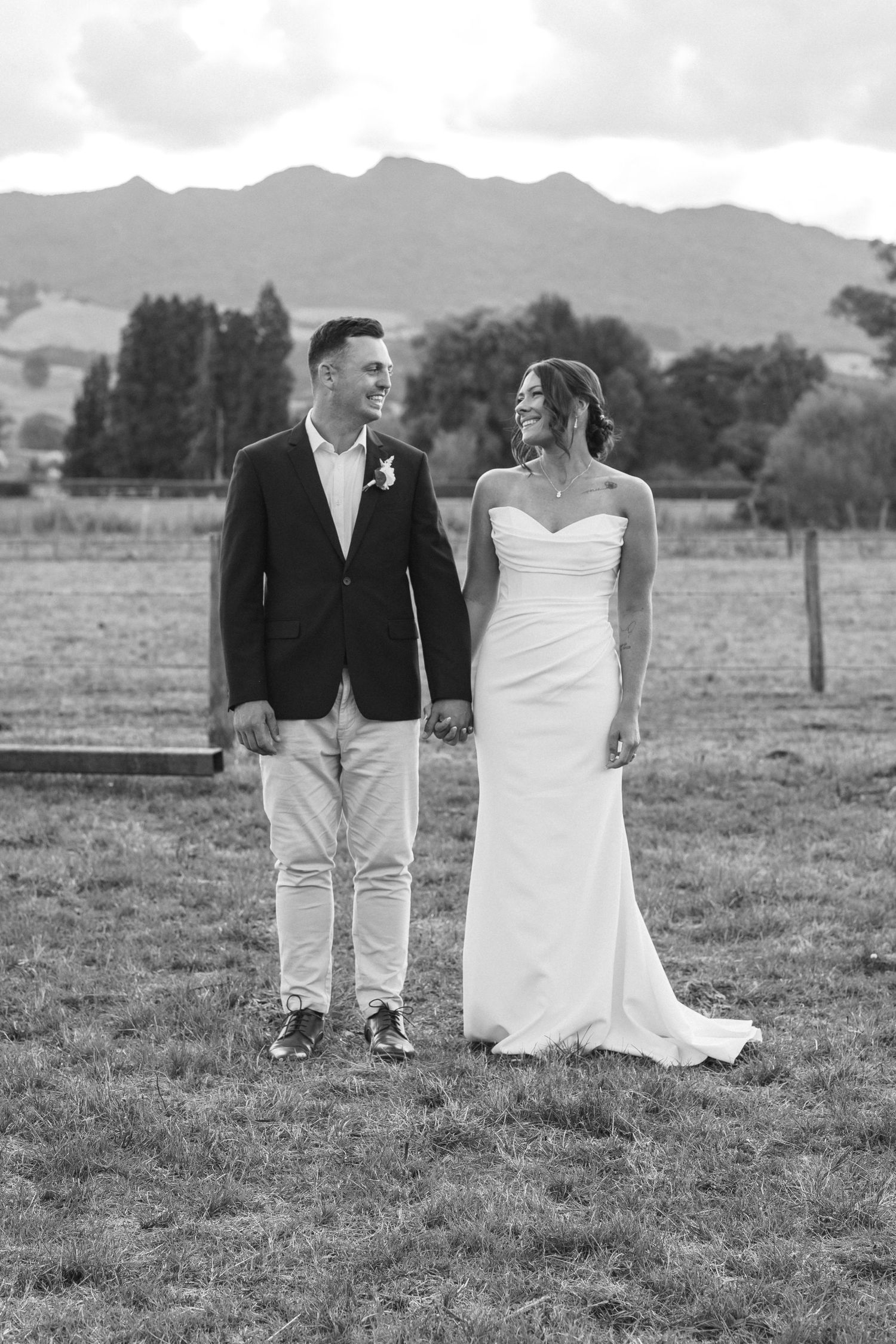 Black and white wedding photo of couple in formal attire standing in field with Pirongia landscape in background.