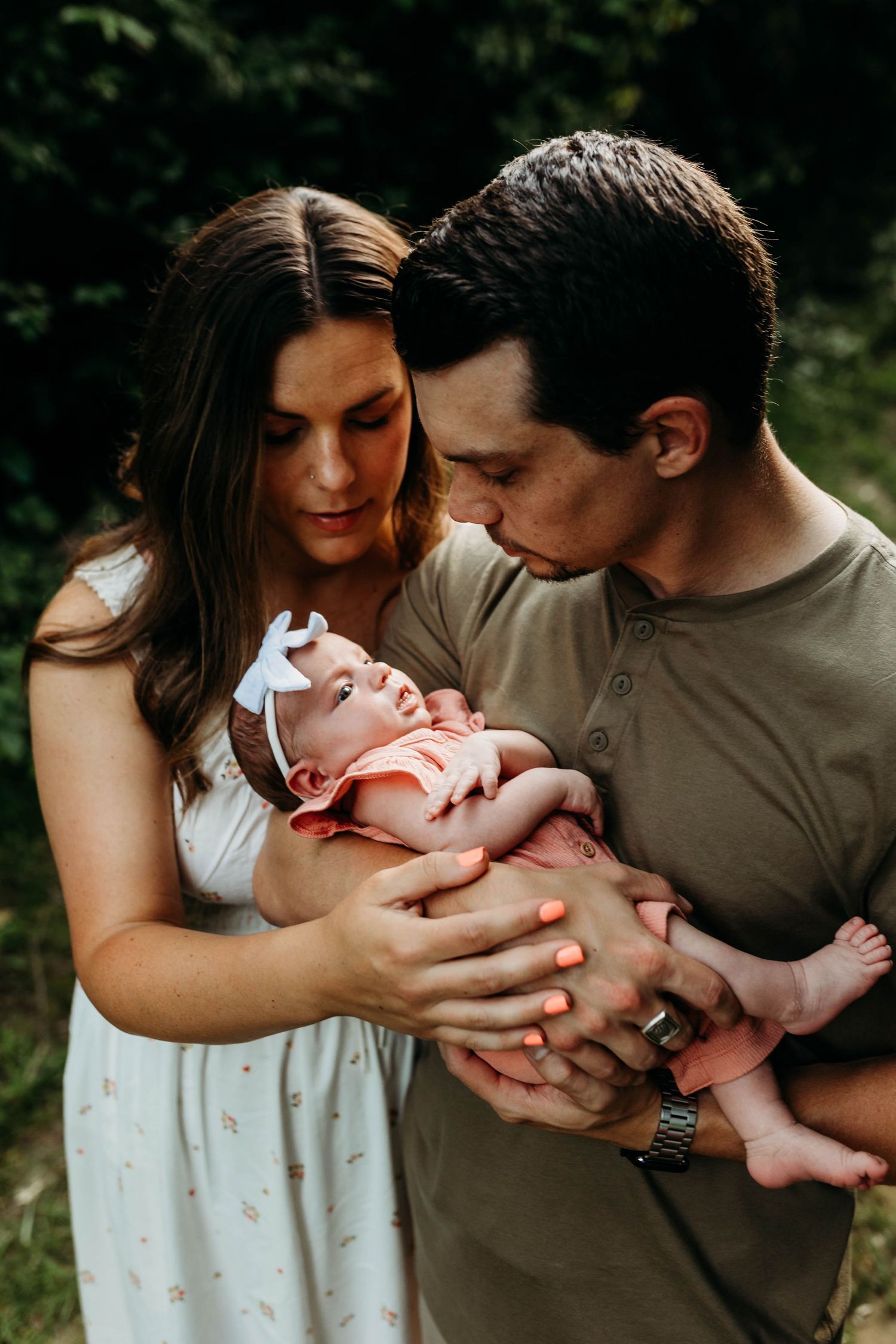 A couple tenderly holds and kisses their newborn baby girl outdoors, with the mother in a white dress.
