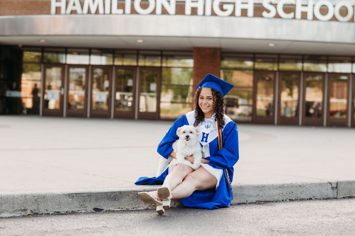High school graduate in blue cap and gown holds white dog outside Hamilton High School.