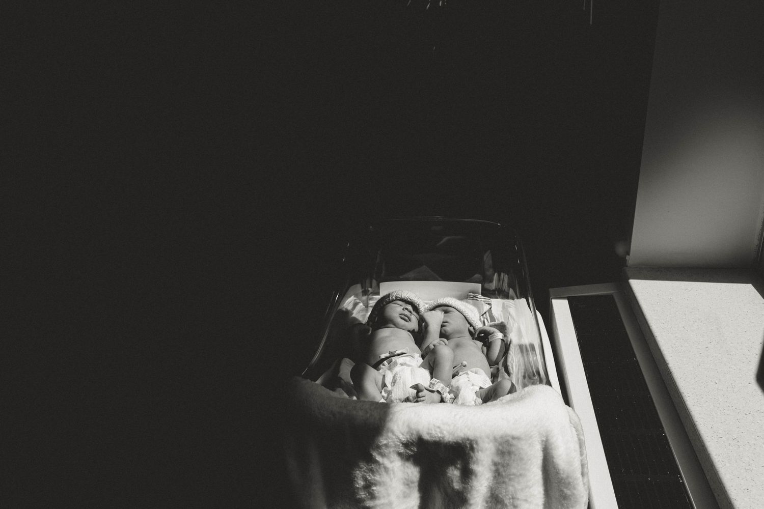 Black and white photo of newborn twins sleeping peacefully side by side in a hospital bassinet.