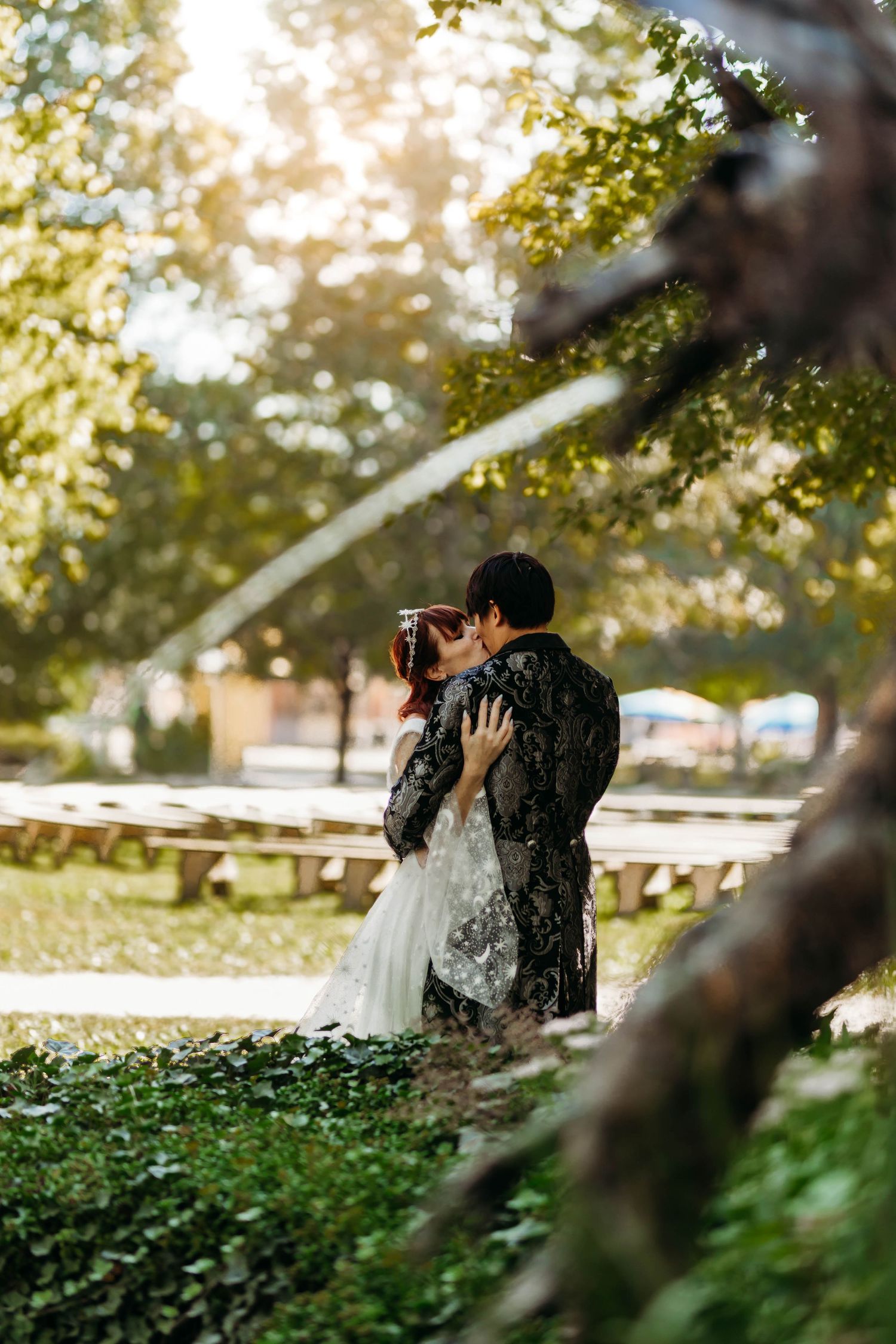 Couple shares a kiss under sunlit green trees in a romantic outdoor wedding setting.