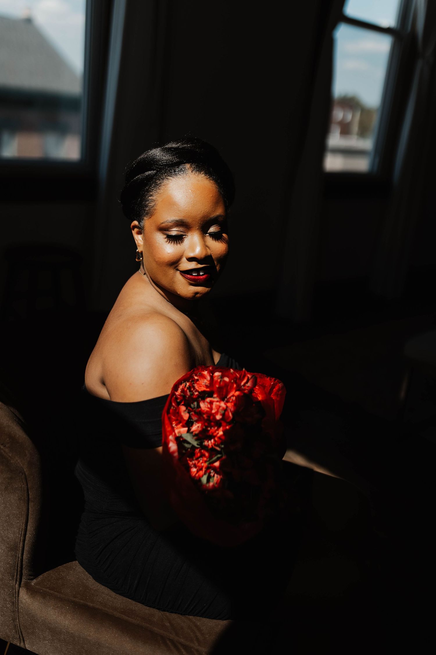A Black woman in a black off-shoulder top holds red flowers, smiling softly in dramatic window light.