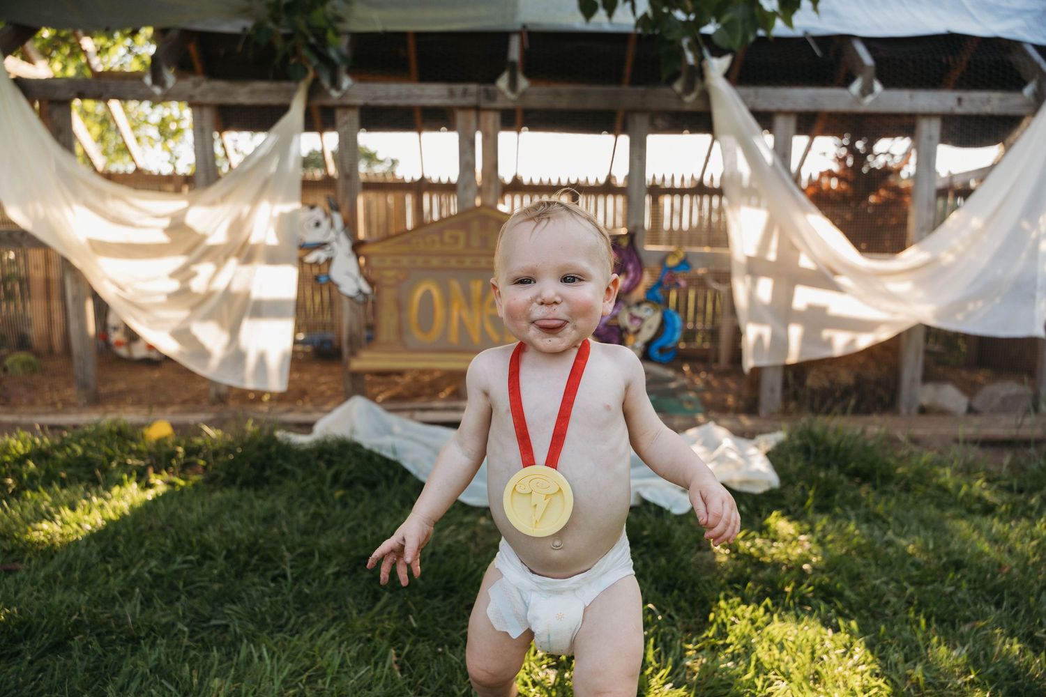 A happy baby in a diaper wearing a gold medal sits on grass outdoors near a rustic wooden structure.