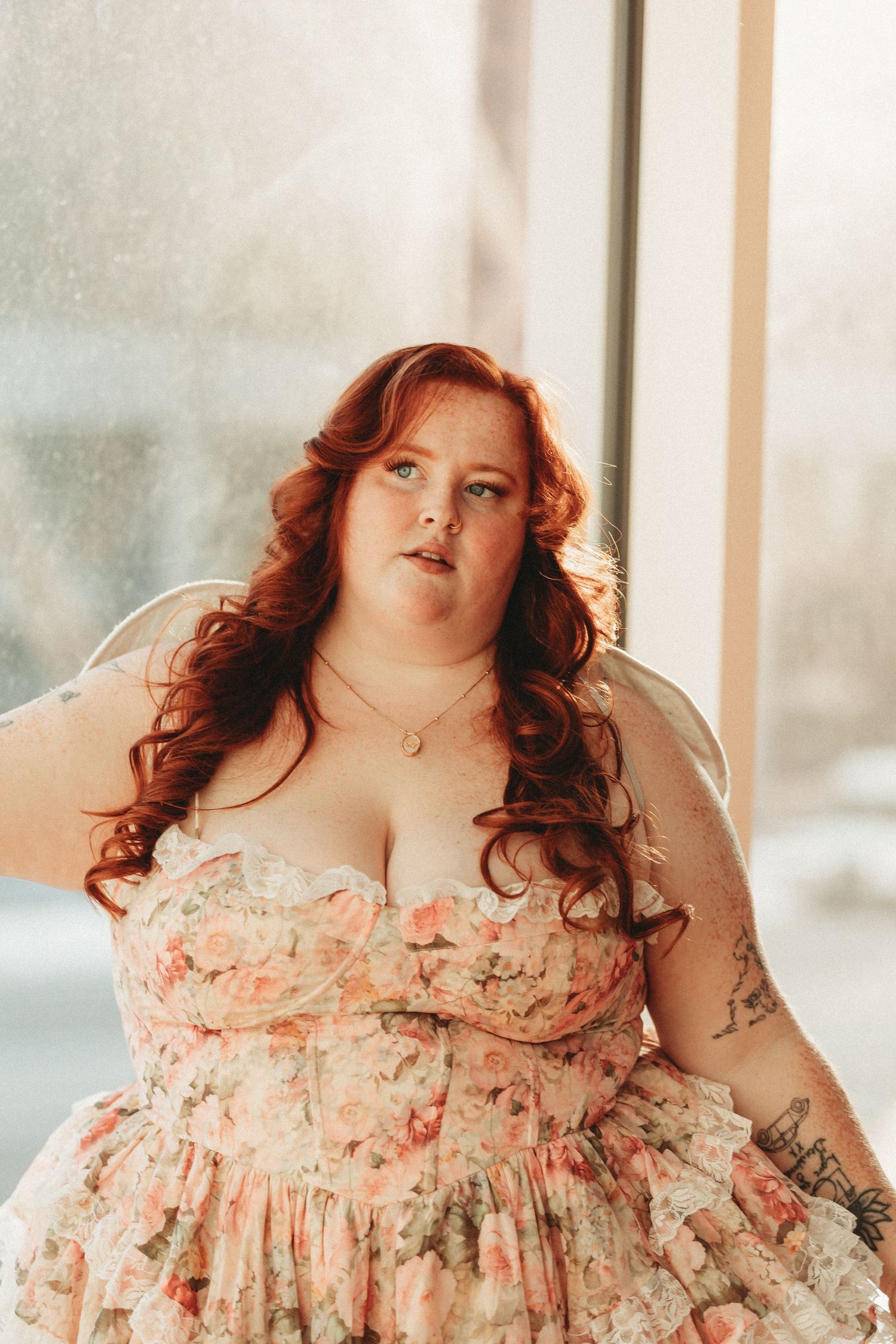 Plus-size woman with red hair poses in floral bustier dress by sunlit window.