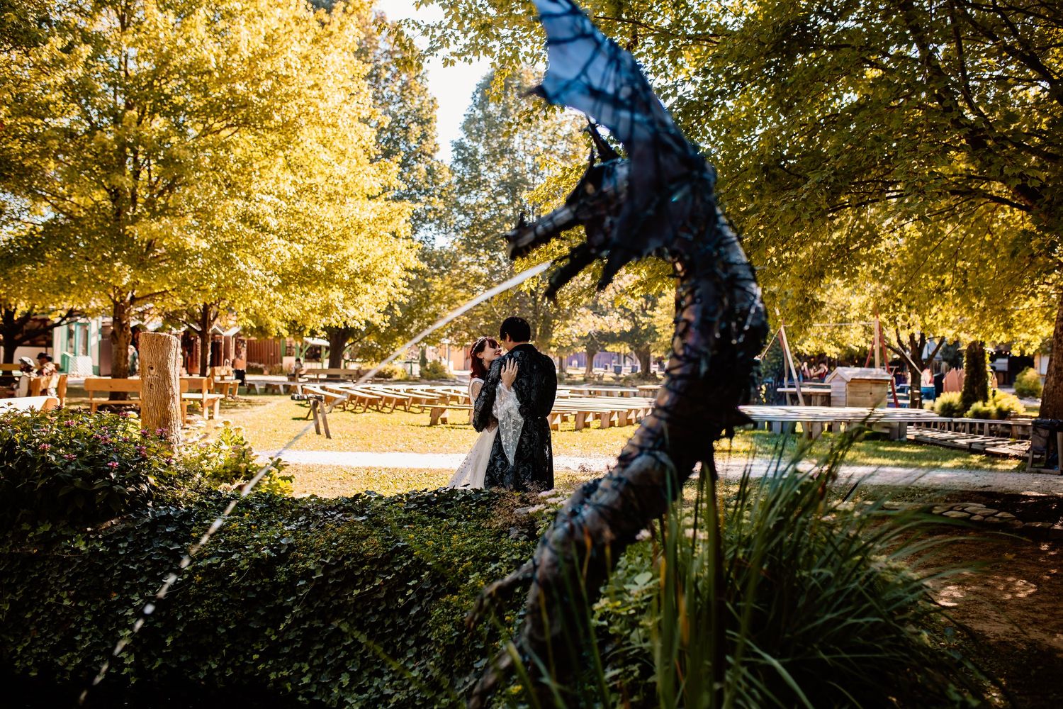 Wedding couple kissing near a blue dragon sculpture fountain surrounded by autumn trees in a park.