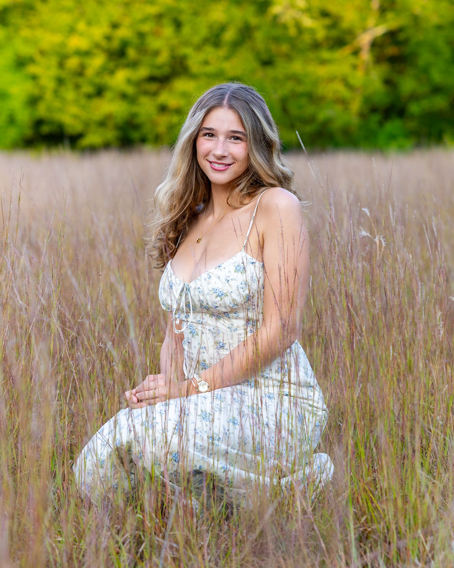 A high school senior in a floral sundress dress poses in tall grass of a golden field during her senior yearbook portrait photography shoot.