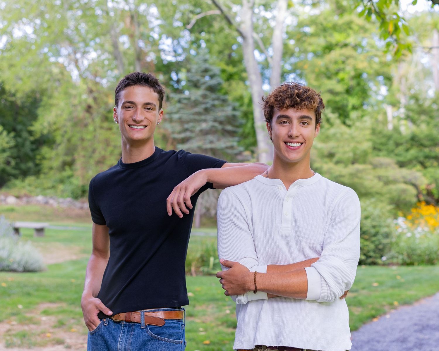 High school senior twins in casual attire pose together outdoors in a park-like setting with trees & greenery in the background in Sudbury.