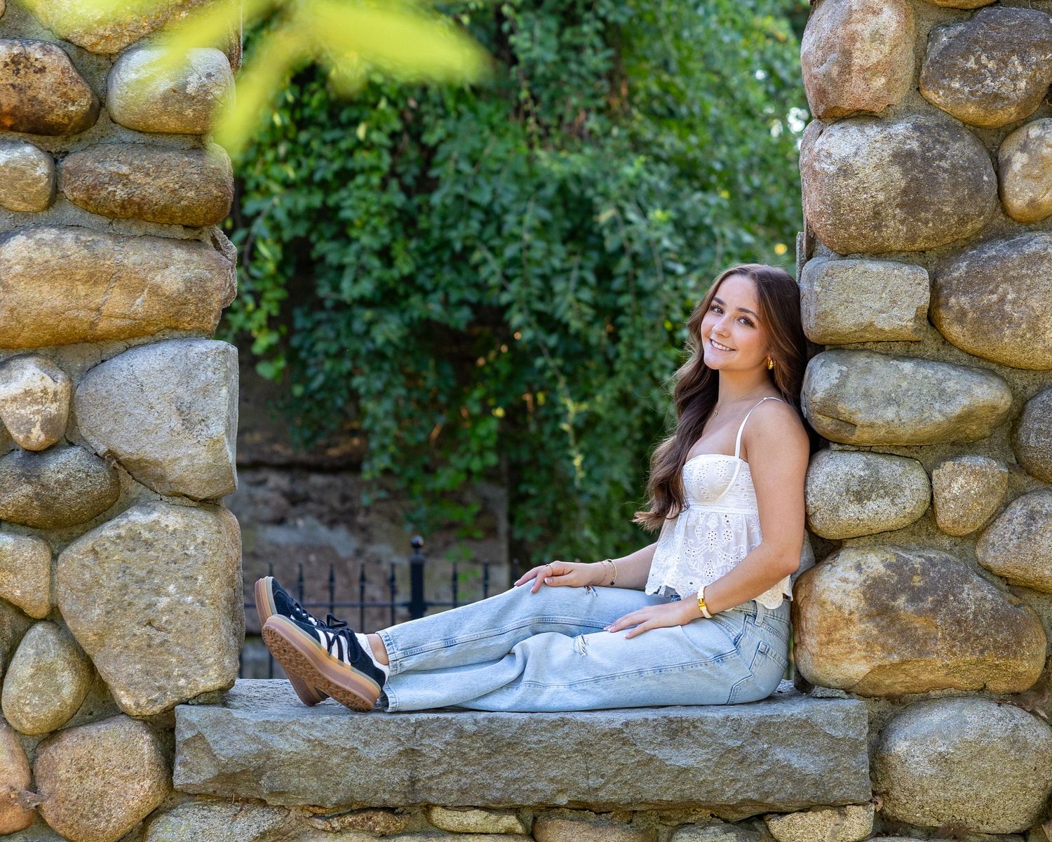 High school senior reclines peacefully on a rustic stone wall of Bancroft Castle Groton, MA surrounded by lush greenery during senior photos