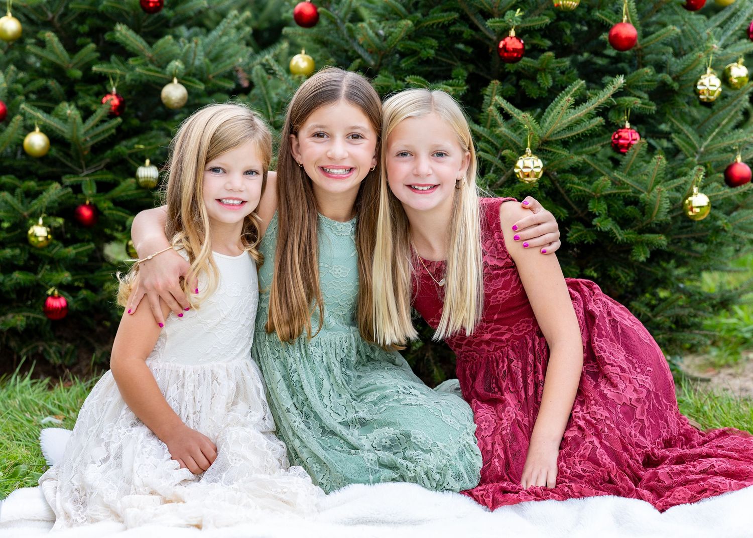 Sisters wearing coordinated red, green & white outfits pose by decorated Christmas tree during their family photos at tree farm in Stow, MA.