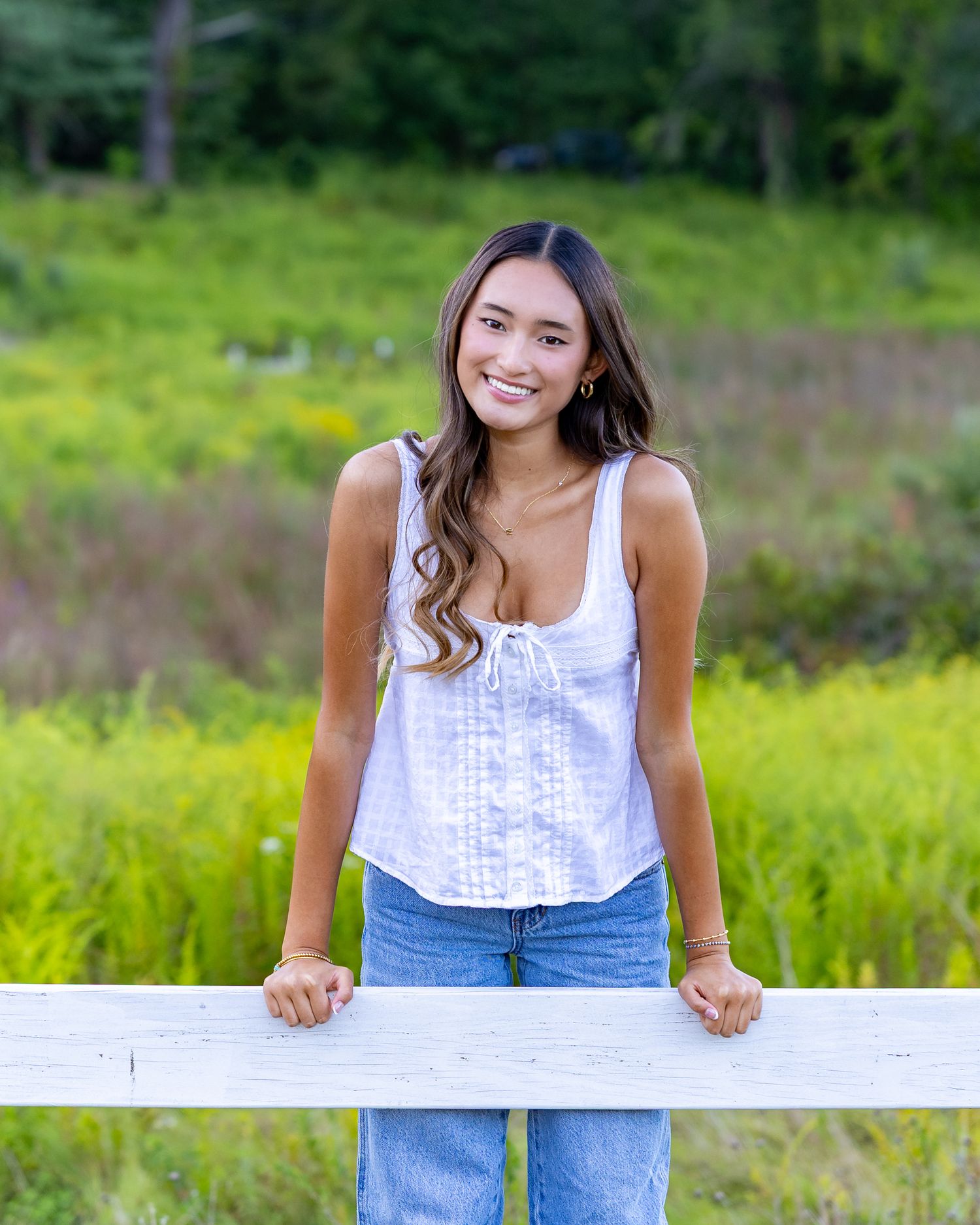 High school senior in a white sleeveless top & jeans sits on a white surface in a lush outdoor setting with greenery during yearbook photos.