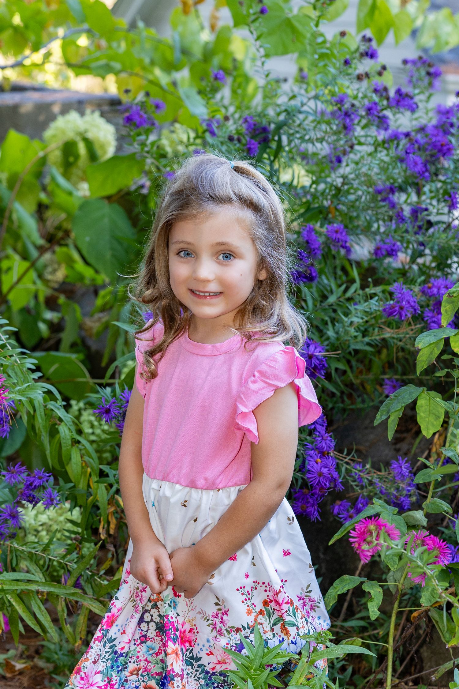 A small child in a pink and white floral dress poses among vibrant purple flowers in a garden setting during her family portraits in Sudbury