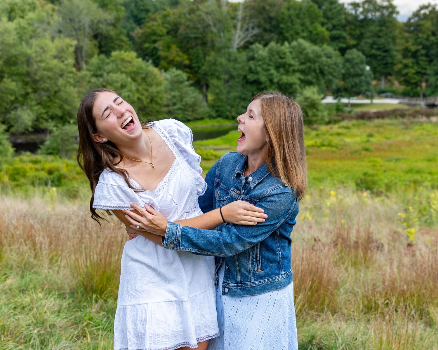 Mother & daughter in white and denim outfits laugh together while posing in a sunny meadow with tall grass at North Bridge in Concord, MA.
