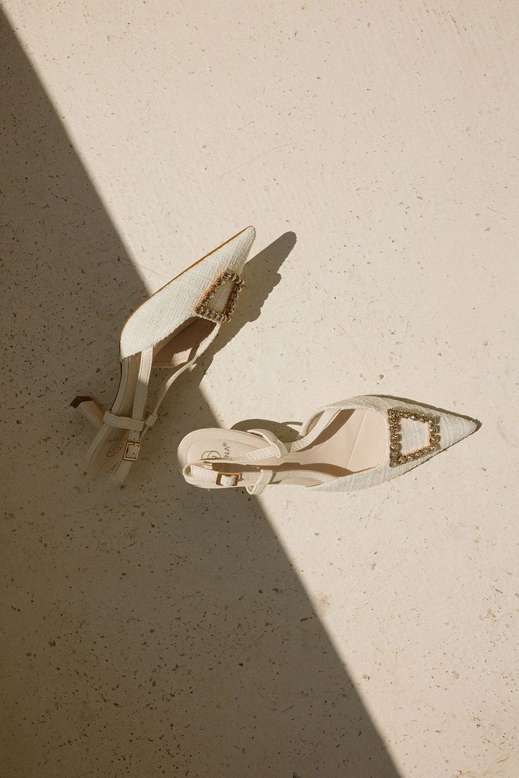 White pointed-toe heels with gold buckle detail casting shadows on a beige stone surface in natural sunlight.
