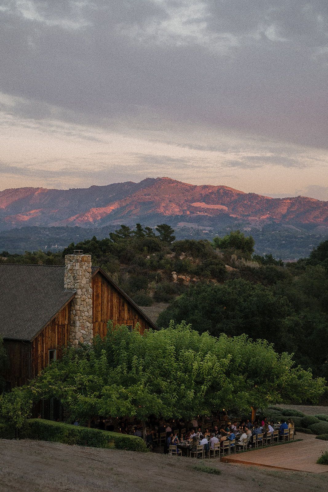 Rustic barn wedding venue at sunset with mountain backdrop and outdoor dining area surrounded by lush greenery.