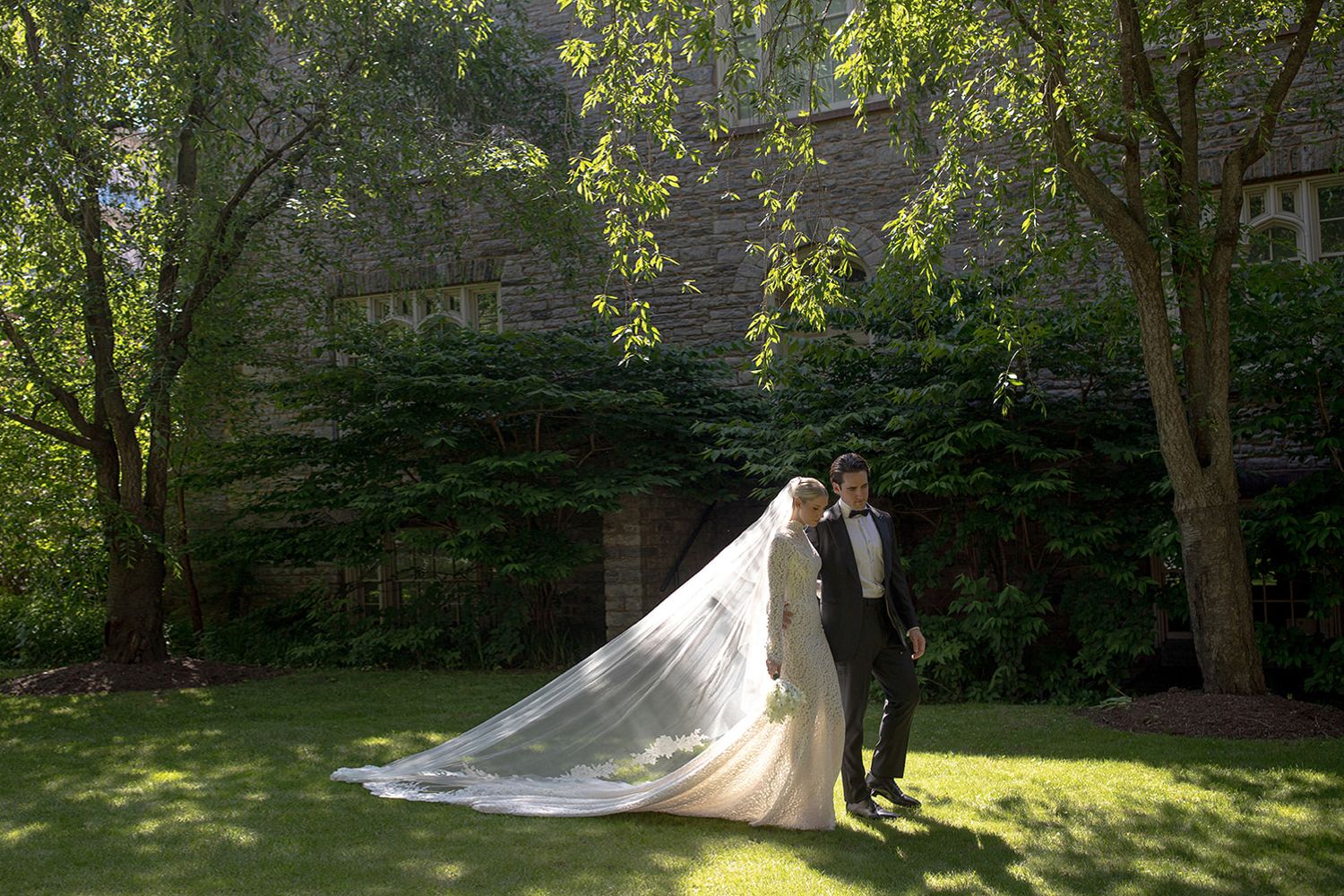 A flowing white wedding dress train extends across a sunlit garden lawn while surrounded by lush green trees.