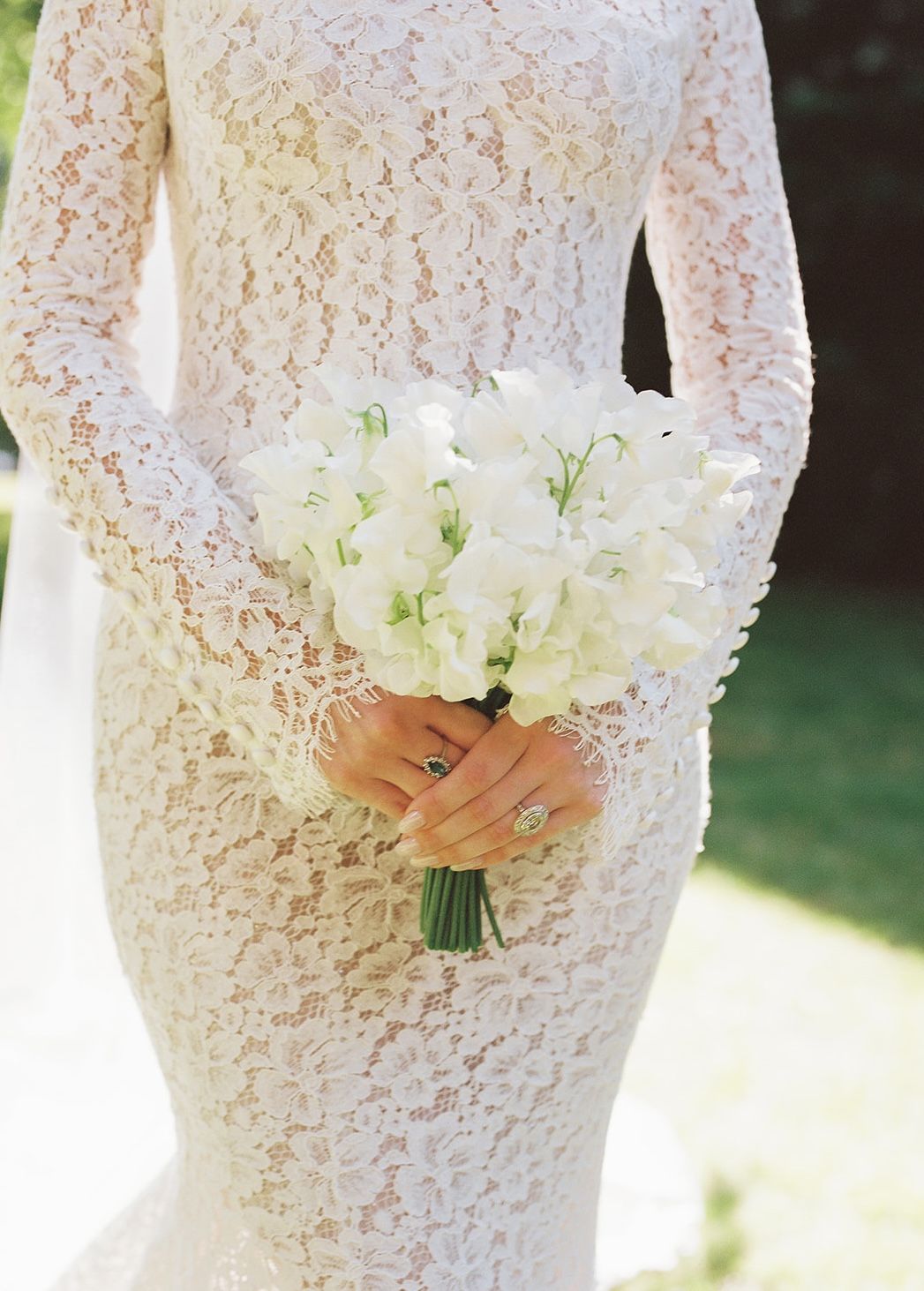 Bride in a long-sleeved ivory lace wedding gown holding a bouquet of white sweet peas with green stems outdoors.