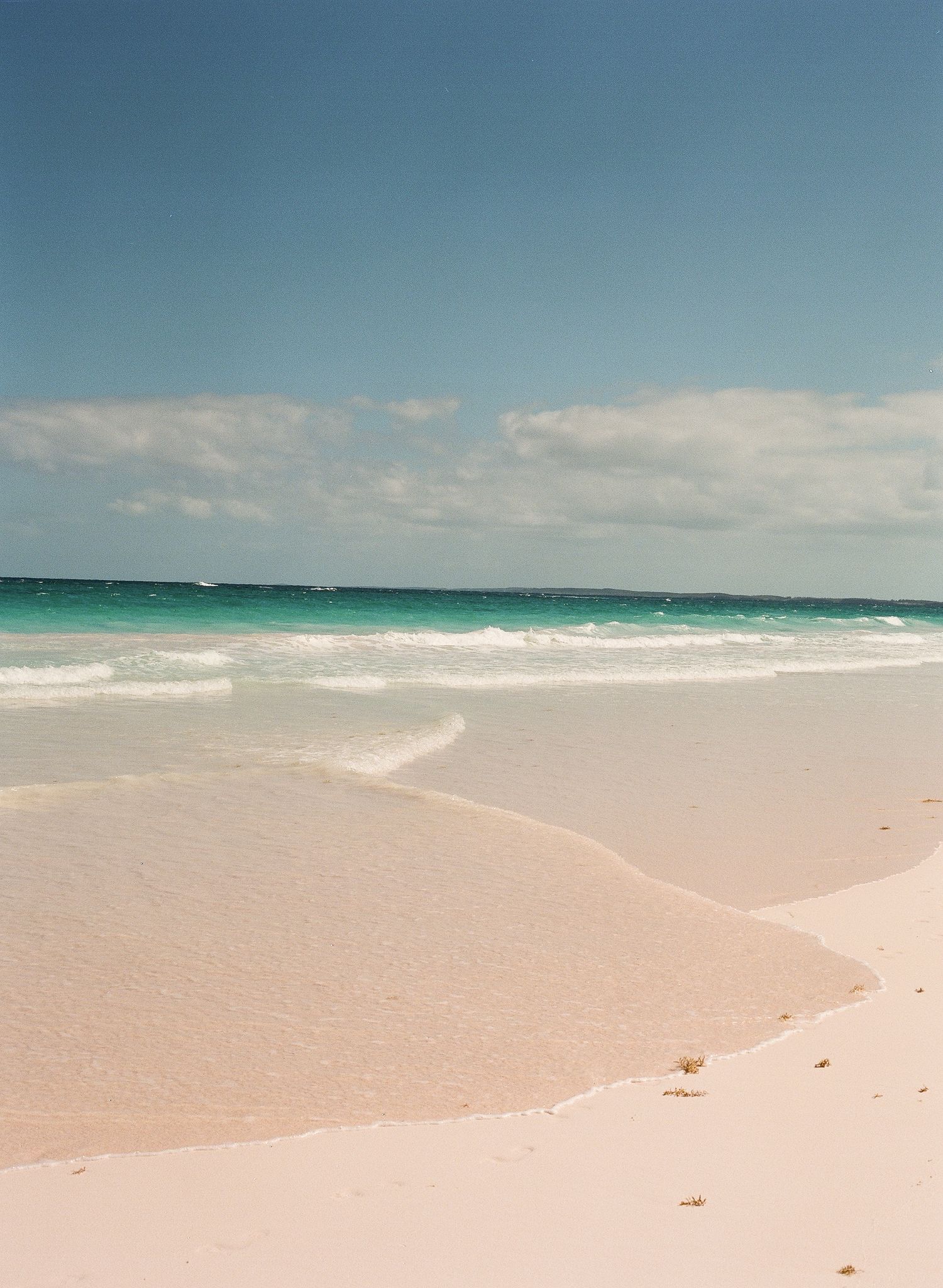 Tranquil panoramic view of pink sandy beach meeting turquoise waters under blue skies with scattered clouds.