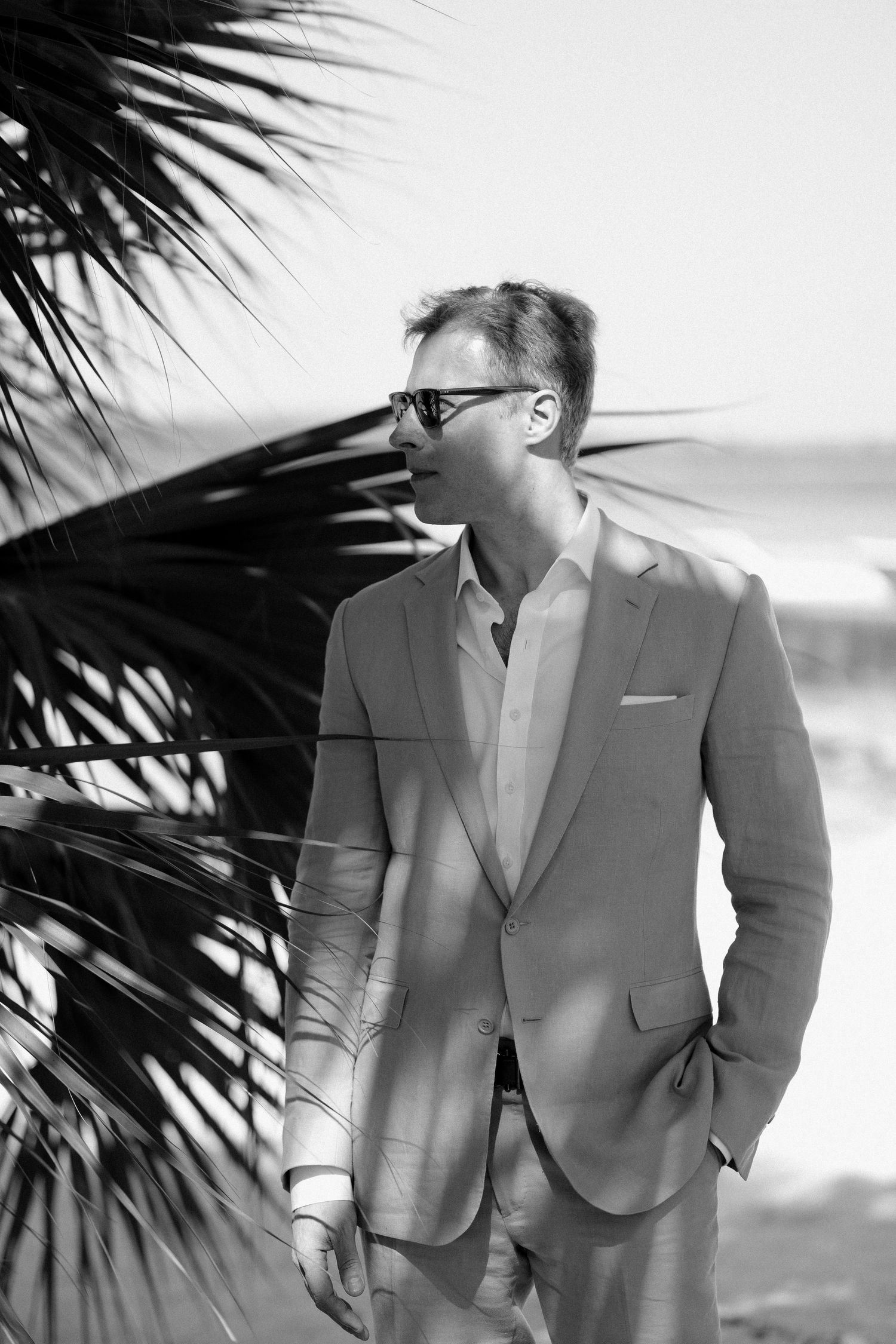 A man in a light gray suit and white shirt poses against a beach backdrop with palm fronds visible.
