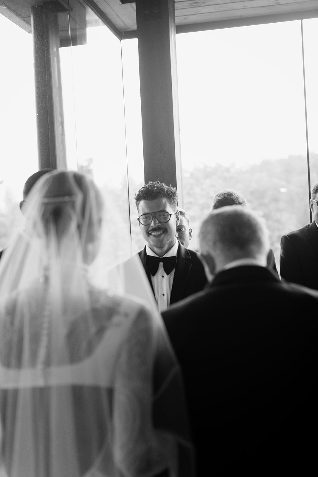 Black and white wedding photo of groom smiling as bride walks down the aisle in elegant veil.