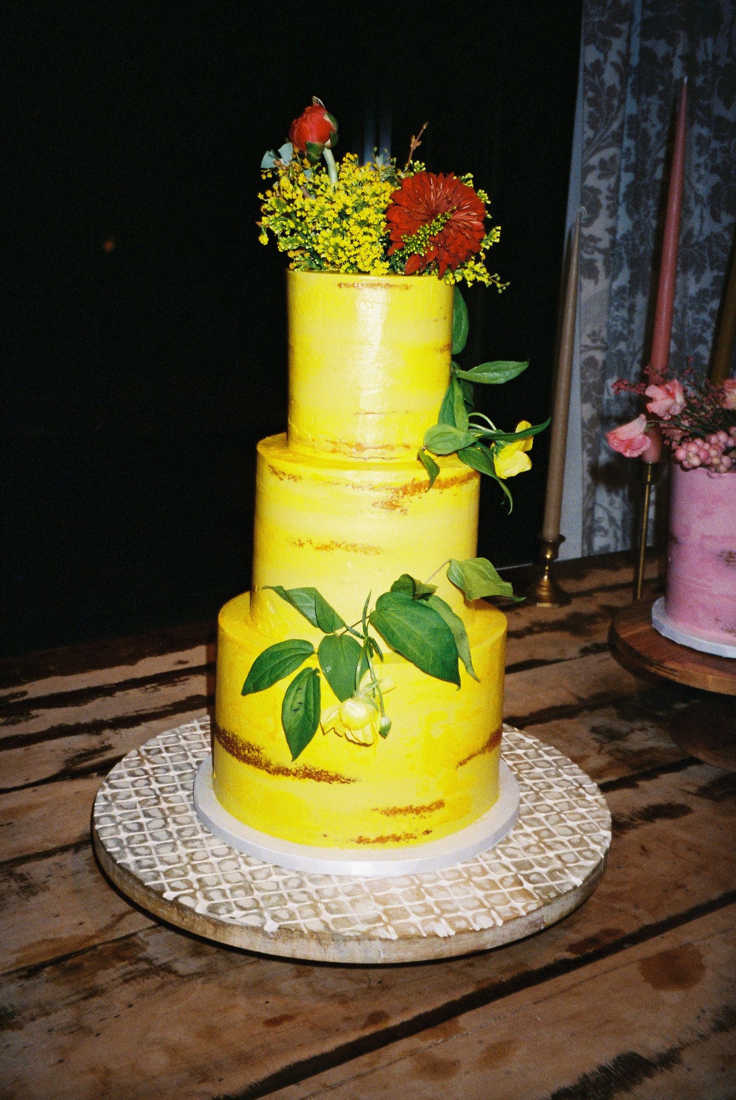 Three-tier yellow semi-naked wedding cake adorned with fresh green leaves, lemons, and colorful floral topper on wooden table.