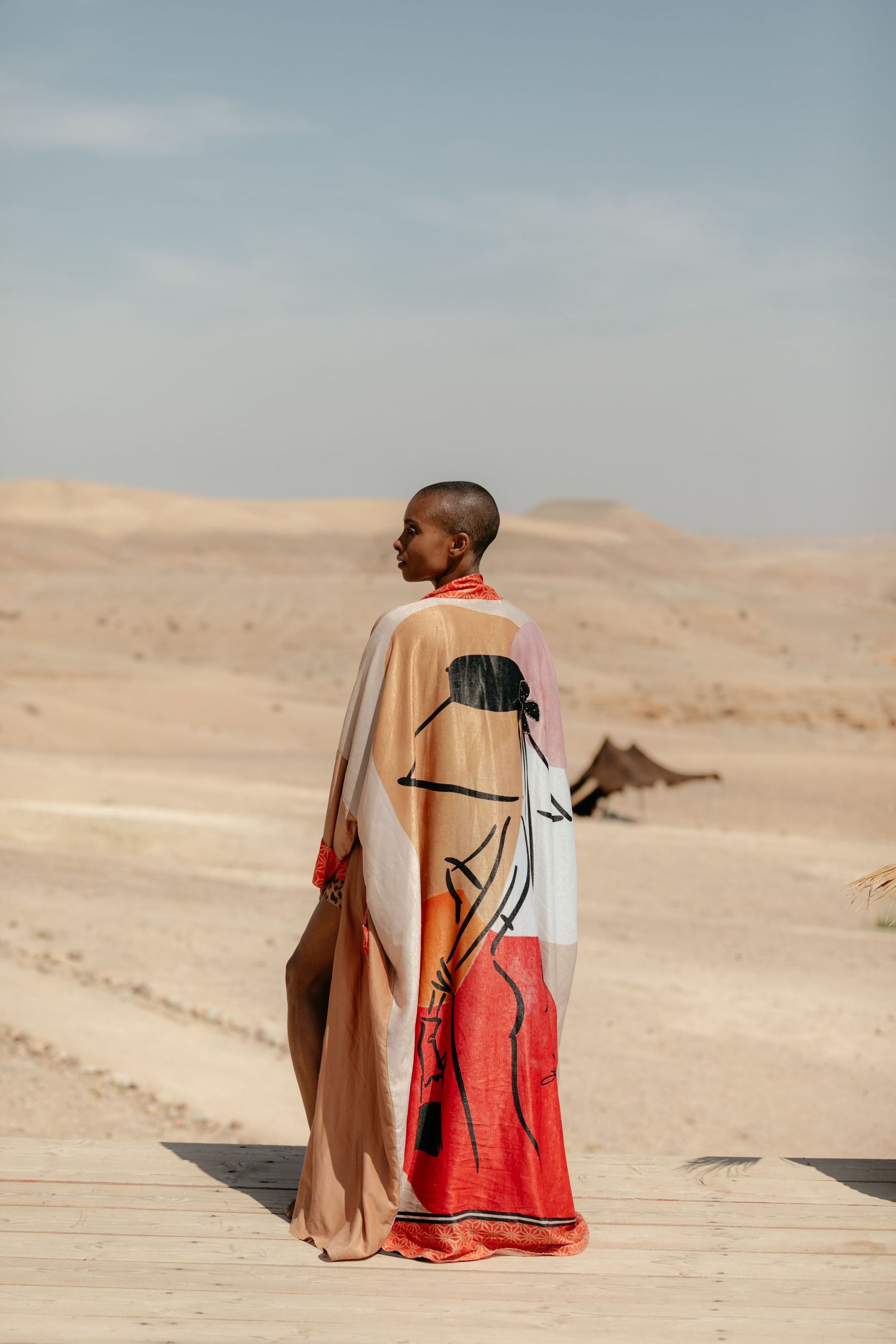 Person in beige and red clothing sitting on desert sand dunes against a pale blue sky.