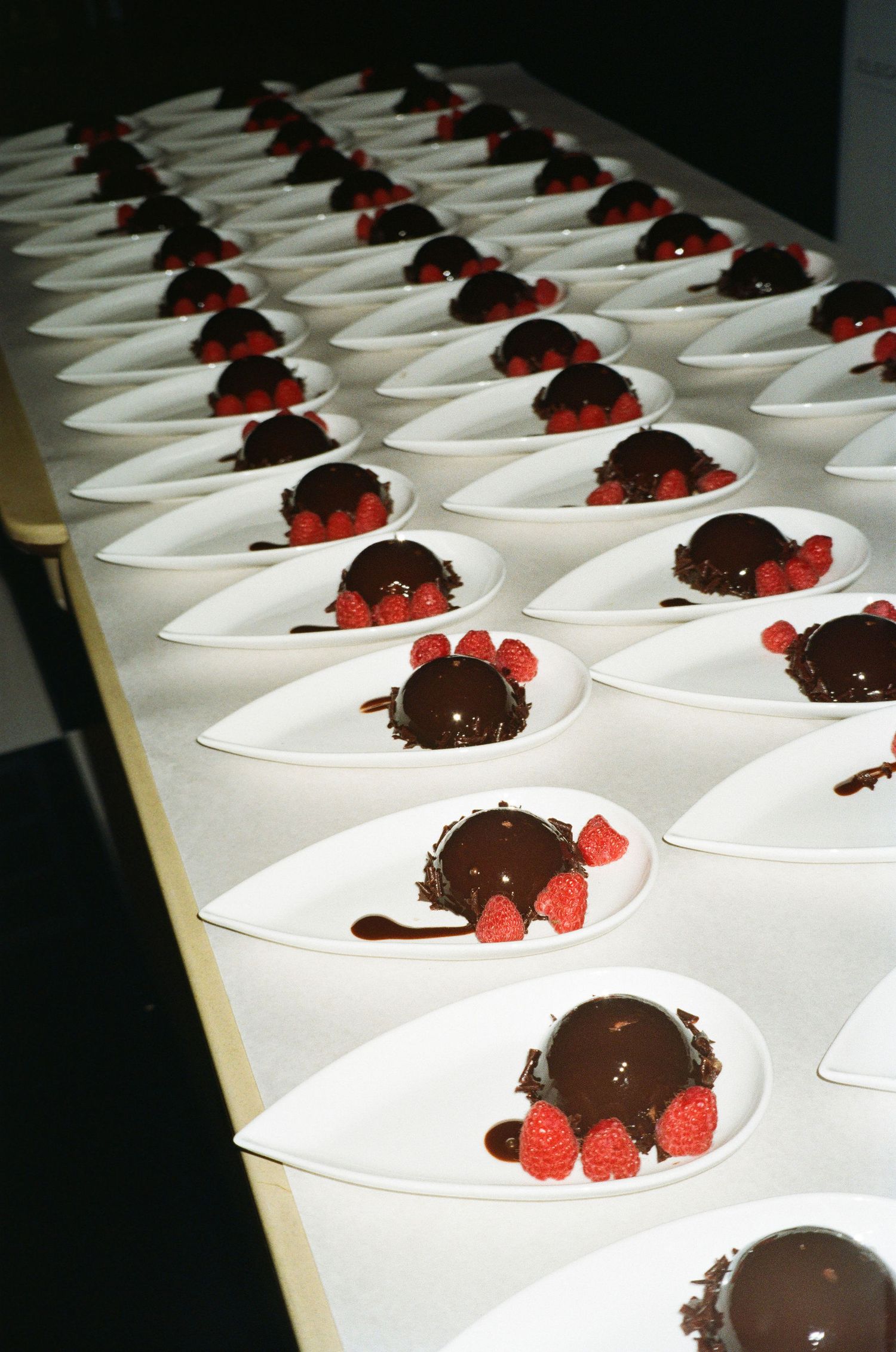 Multiple white plates arranged in rows featuring chocolate desserts garnished with fresh red raspberries.