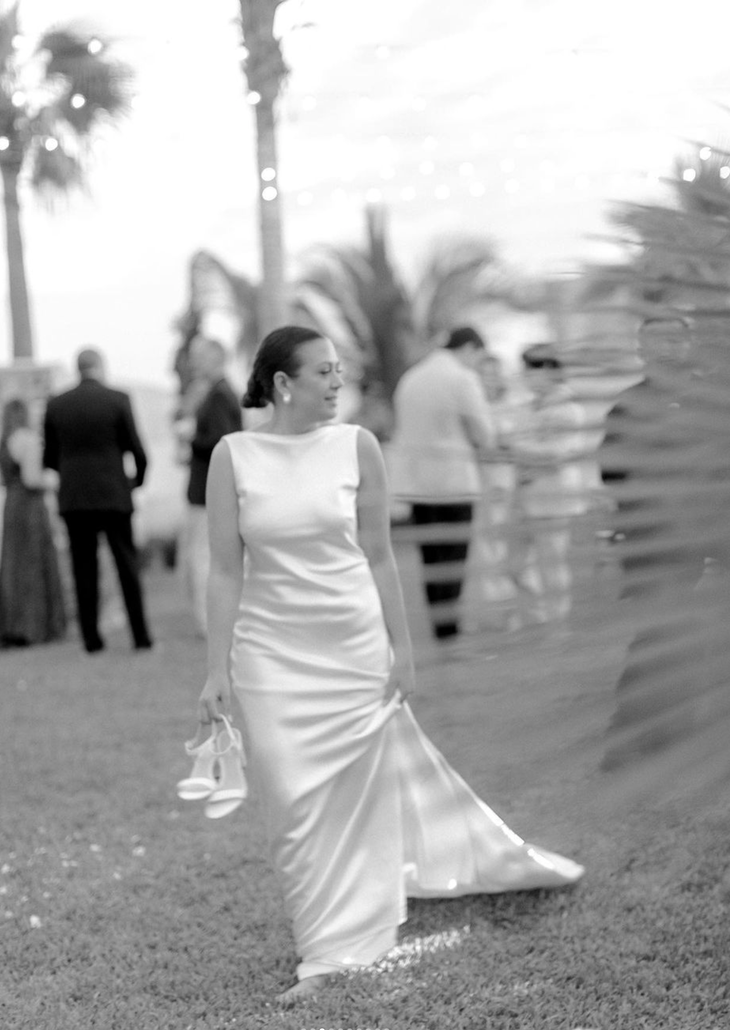 Black and white photo of a bride in sleek white dress walking on grass with palm trees in background.