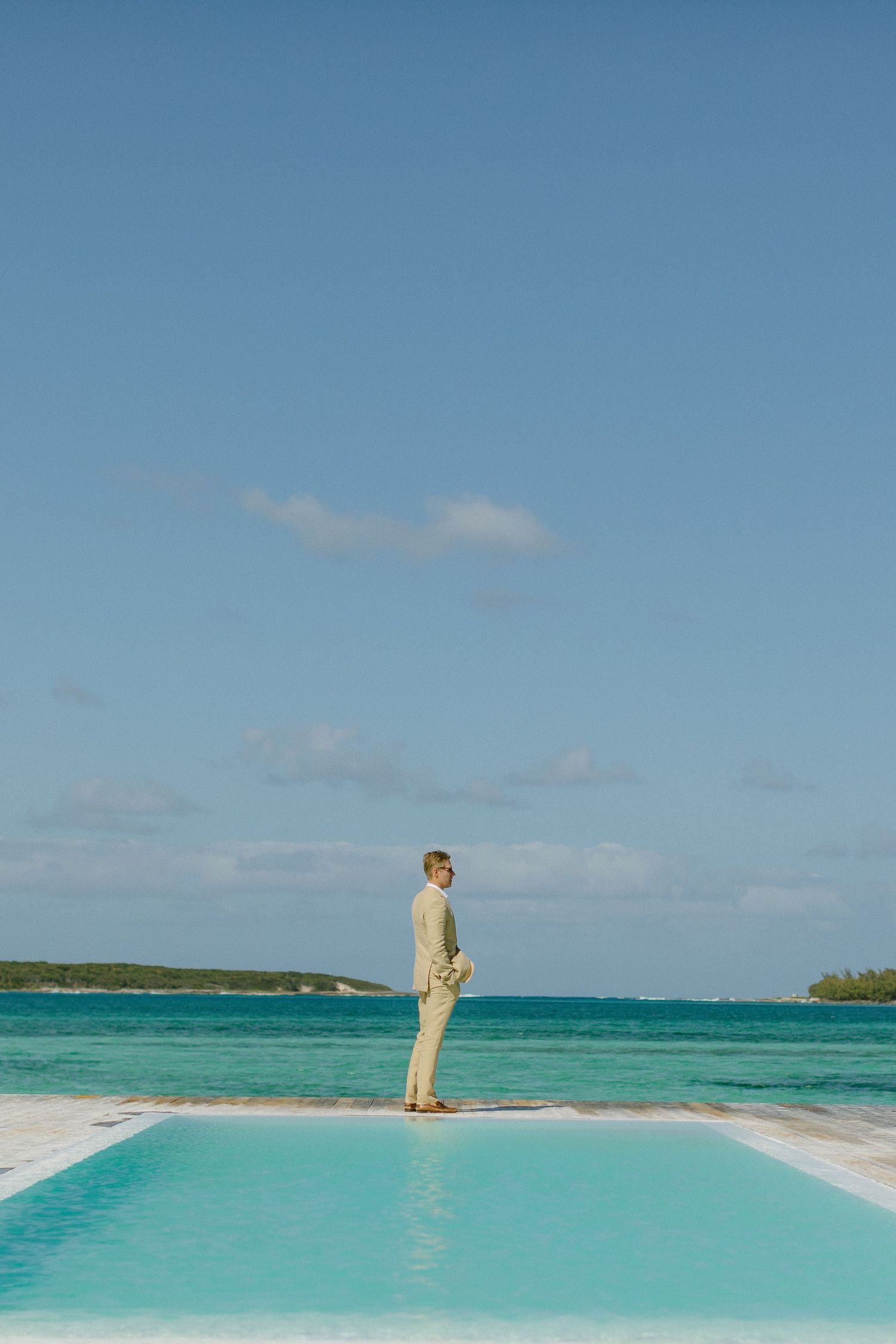 Person in light suit standing on edge of infinity pool overlooking turquoise ocean.