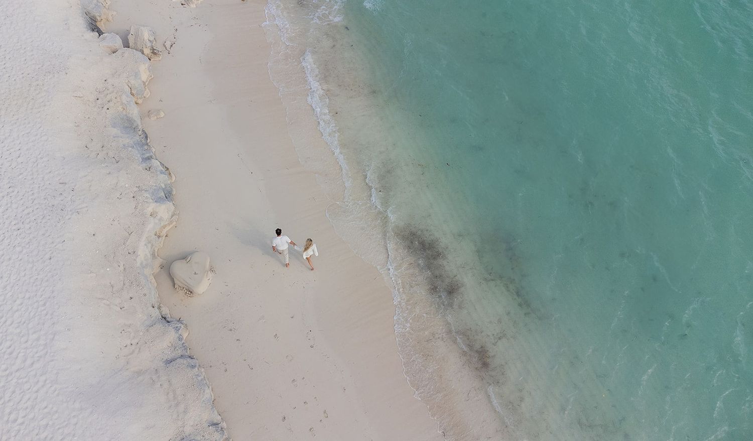 Aerial view of two people relaxing on a white sandy beach beside crystal clear turquoise ocean water.
