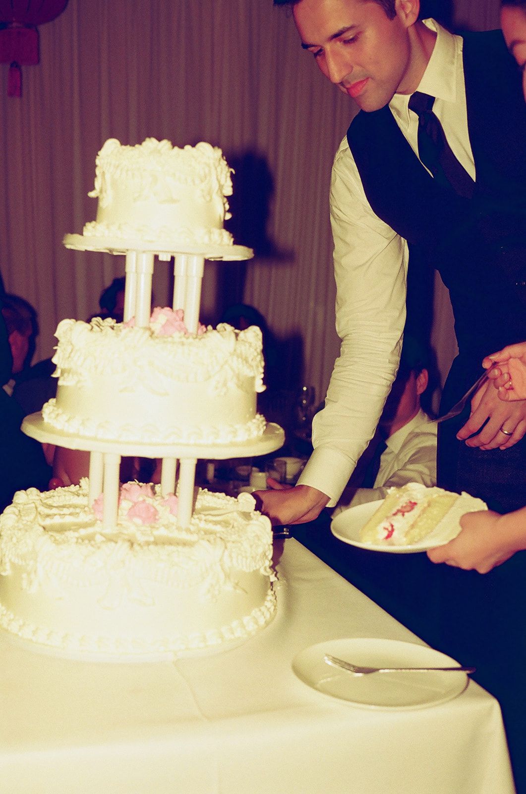 Groom in black suit cutting a three-tier white wedding cake with pink roses at a reception.