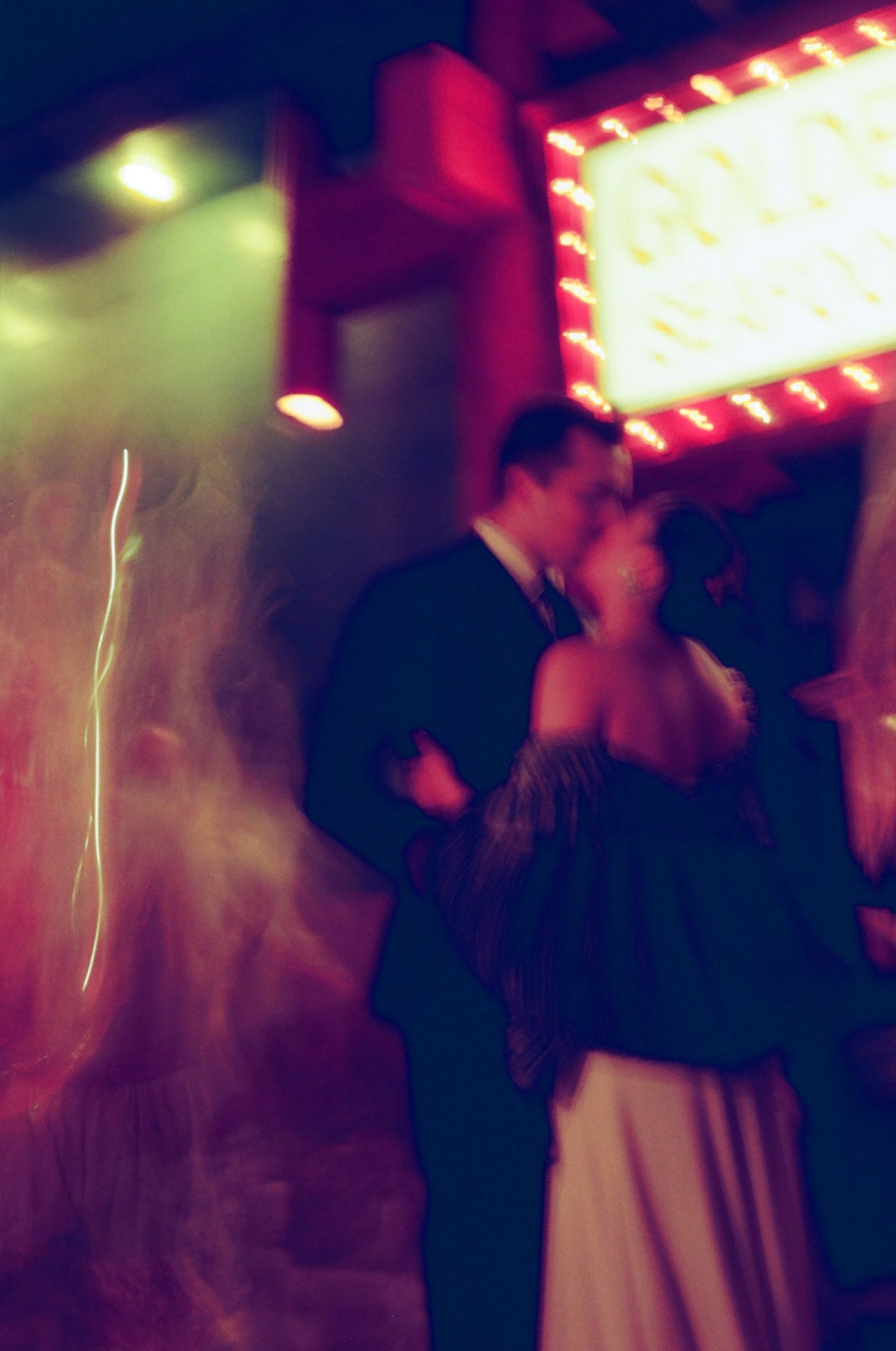Blurry, dreamy night photo of a couple in formal attire kissing beneath a glowing marquee sign with red and orange lights.