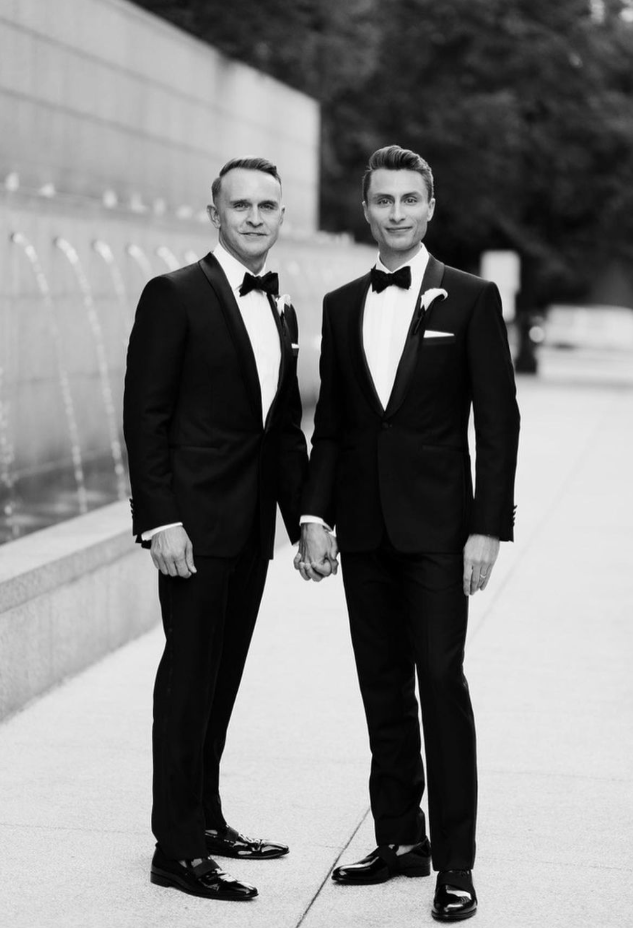 Two people in classic black tuxedos with bow ties pose together at a formal wedding ceremony.