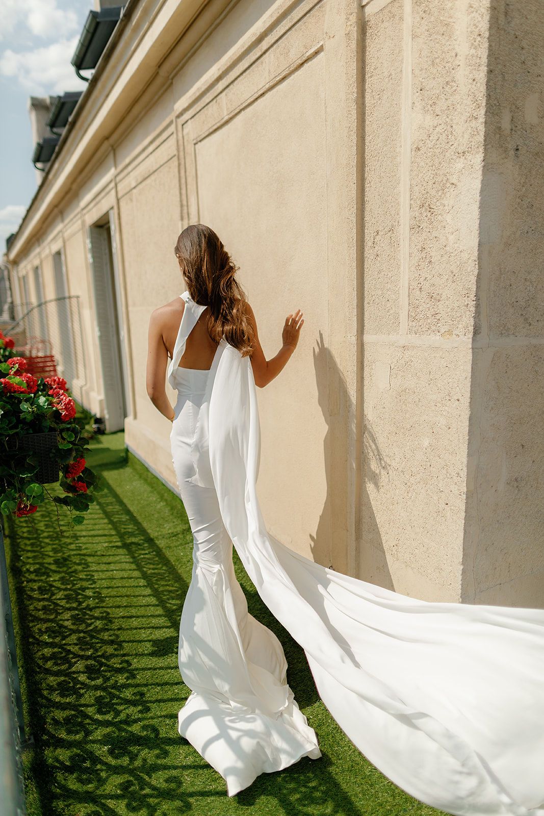 Bride in elegant white gown with flowing train stands against beige stone wall in lush garden.