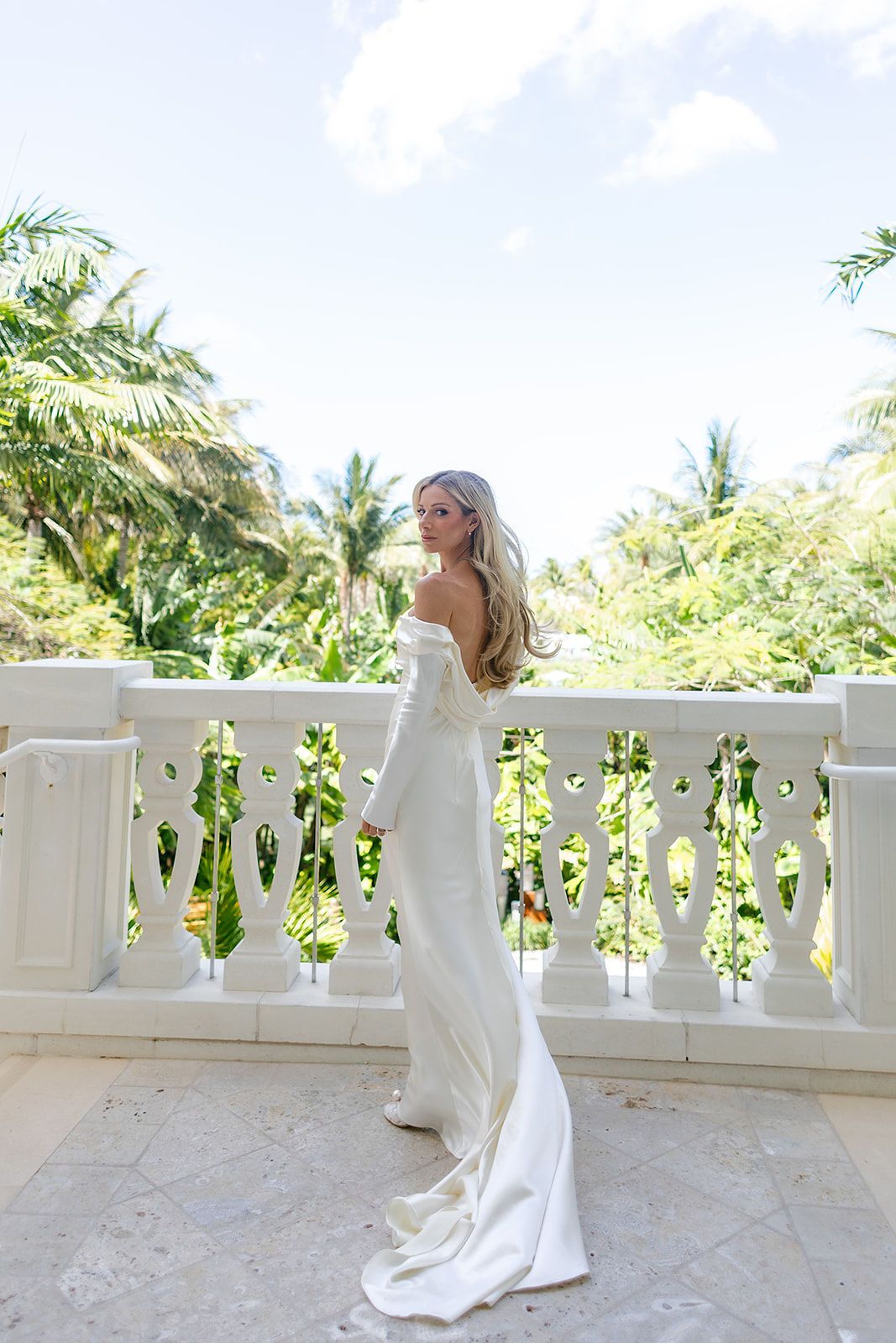 Bride in elegant off-shoulder white gown on tropical balcony with lush palm trees and white balustrade.