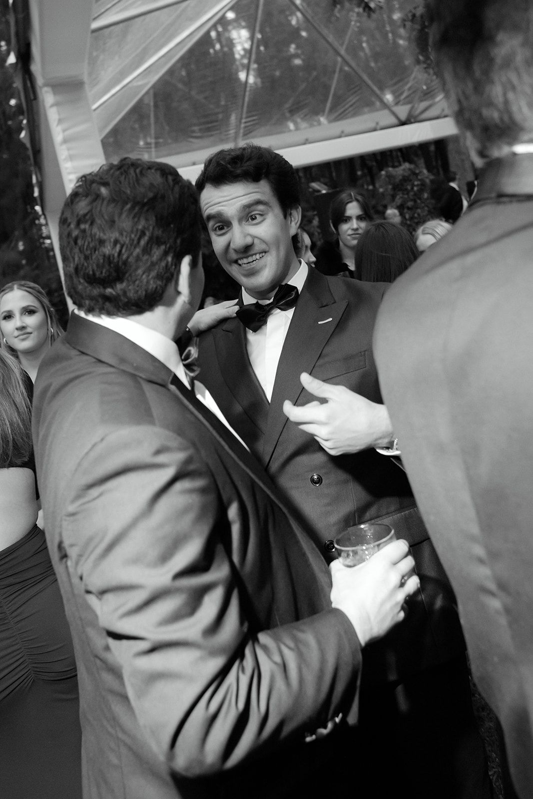 A crowded formal event scene with people in dark suits mingling under dim lighting.